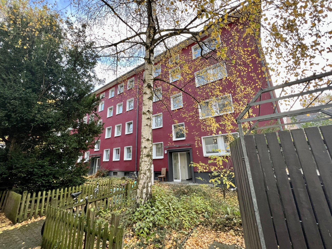 Rotes Wohngebäude mit mehreren Fenstern, Bäumen und Holzzaun im Vorgarten.