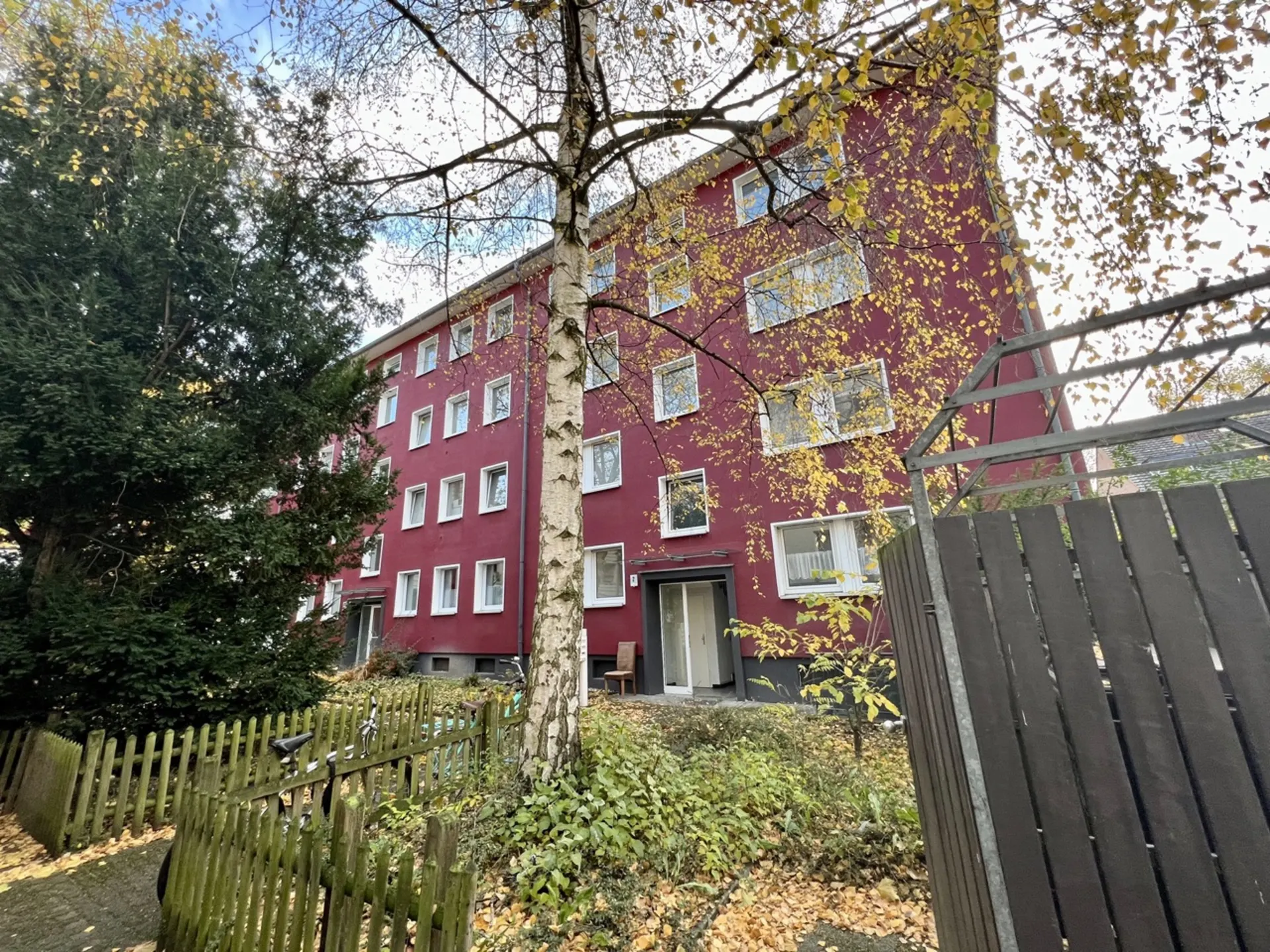 Rotes Wohngebäude mit mehreren Fenstern, Bäumen und Holzzaun im Vorgarten.