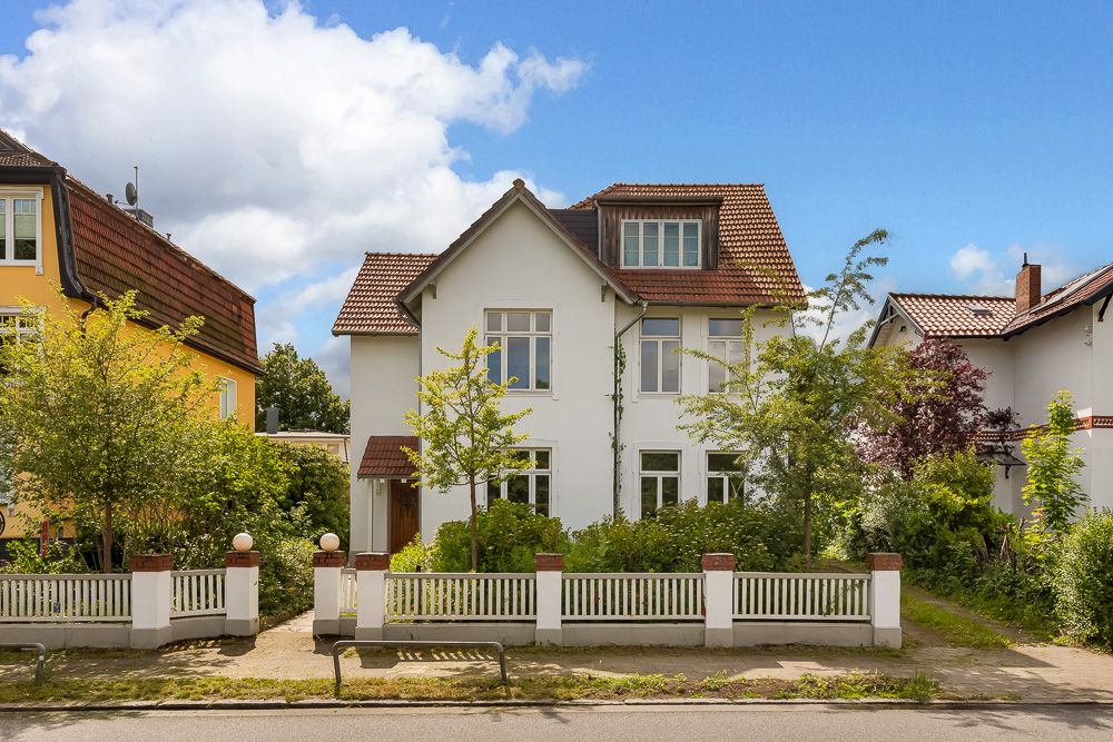 Zweistöckiges weißes Wohnhaus mit Garten und Zaun, Bäume davor, blauer Himmel im Hintergrund.