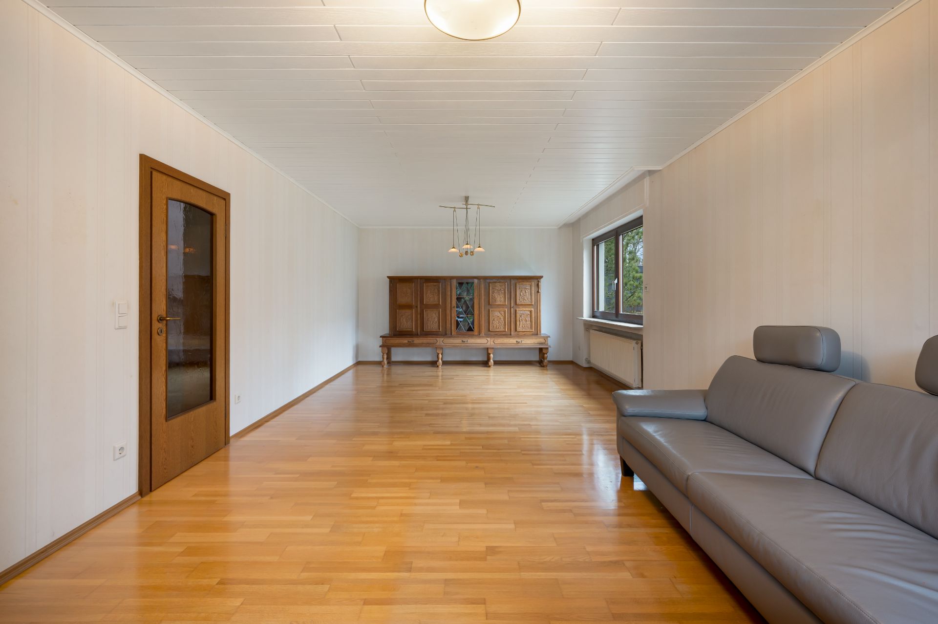 Helles Wohnzimmer mit Holzfußboden, grauem Sofa, großem Fenster und Holzschrank.