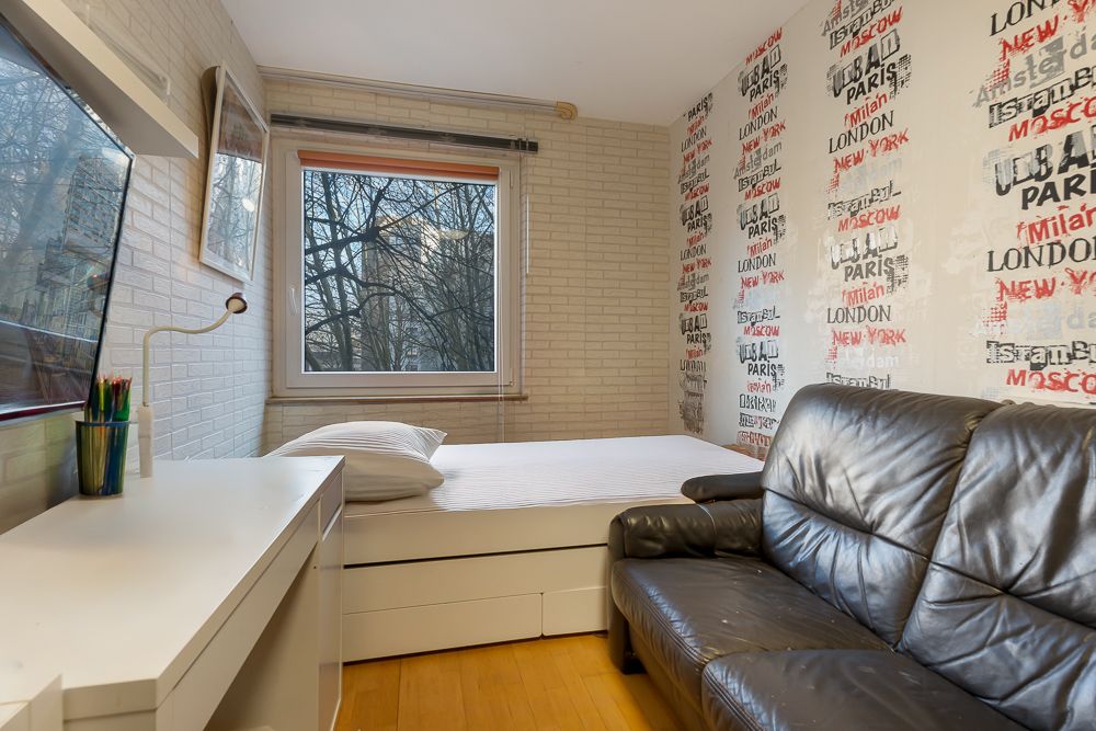 Kleines Zimmer mit Bett, Schreibtisch, schwarzem Sofa und Fenster mit Blick auf Bäume.