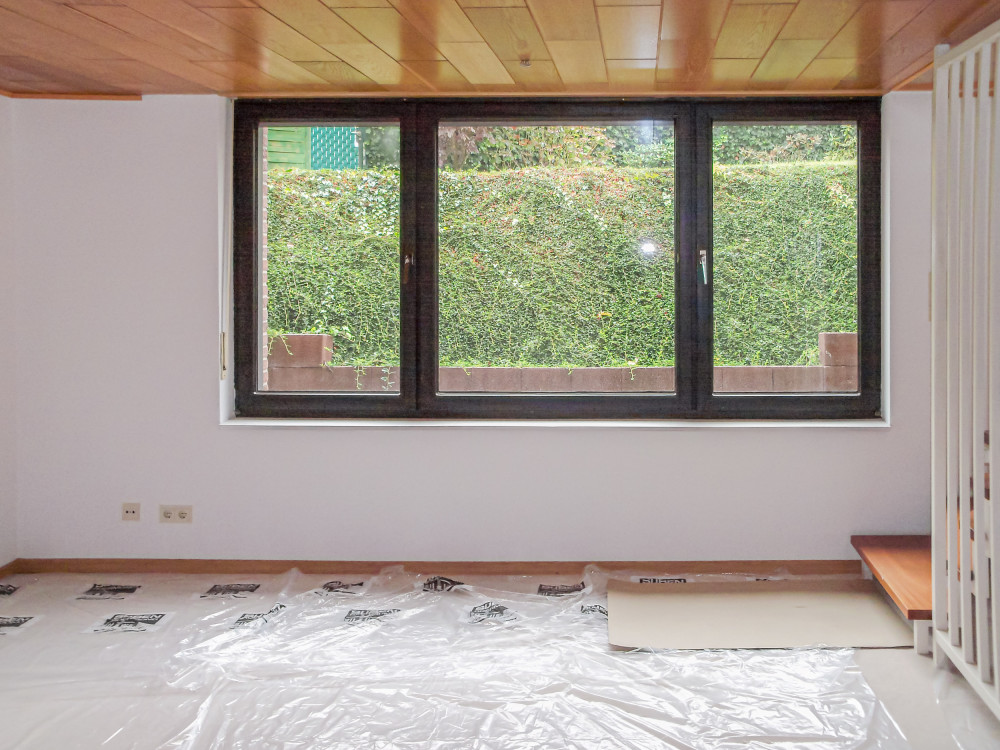Helles Zimmer mit großem Fenster, Blick auf grüne Hecke, Kunststofffolie auf dem Boden.