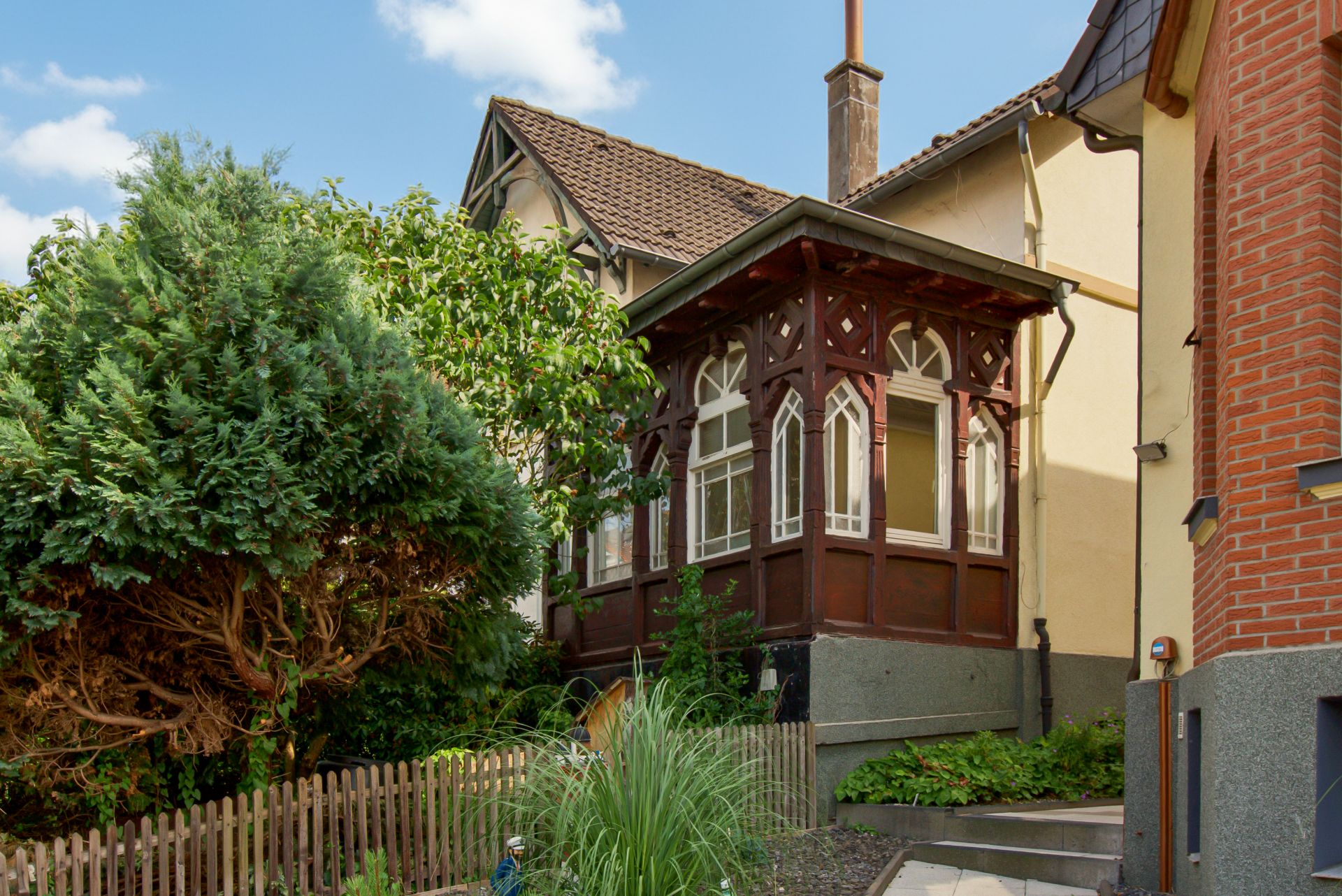 Wohnhaus mit verglastem Erker, Holzzaun und bewachsenem Gartenbereich im Vordergrund.