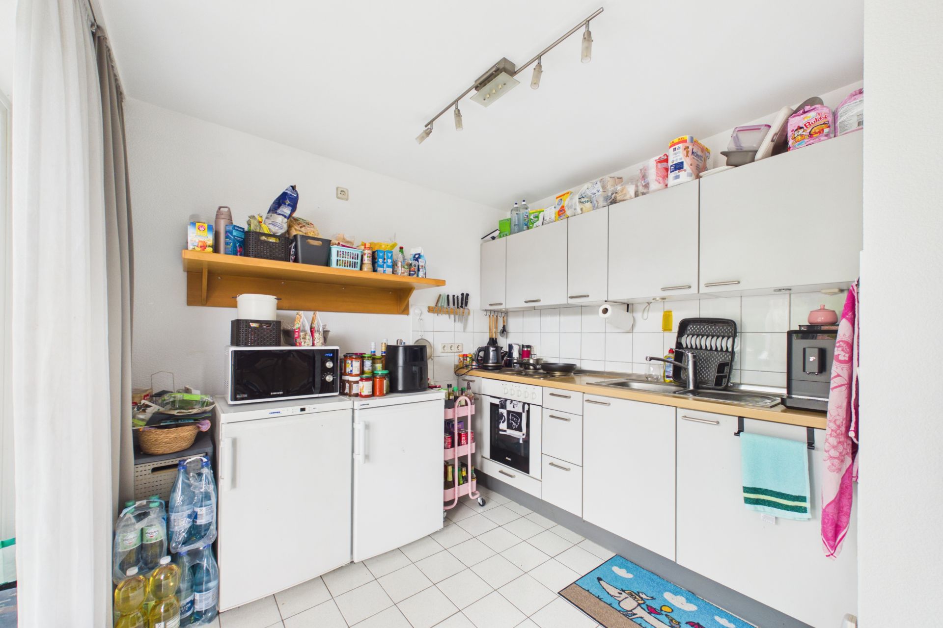Helle Küche mit weißen Schränken, Kühlschrank, Mikrowelle und Küchenutensilien auf den Arbeitsflächen.