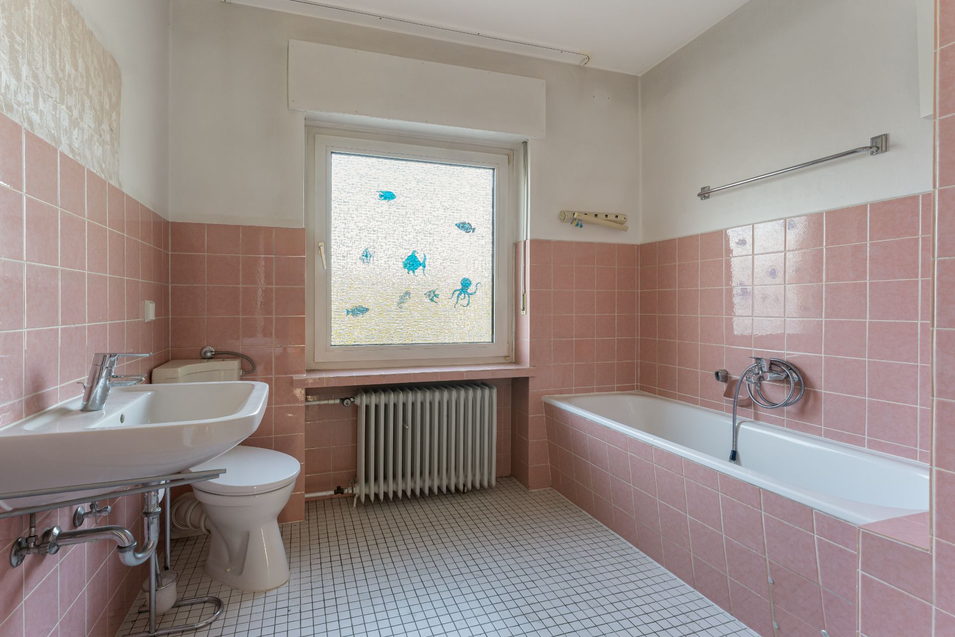 Badezimmer mit rosa Fliesen, Badewanne, Waschbecken, Toilette und Fenster mit Fischmotiven.