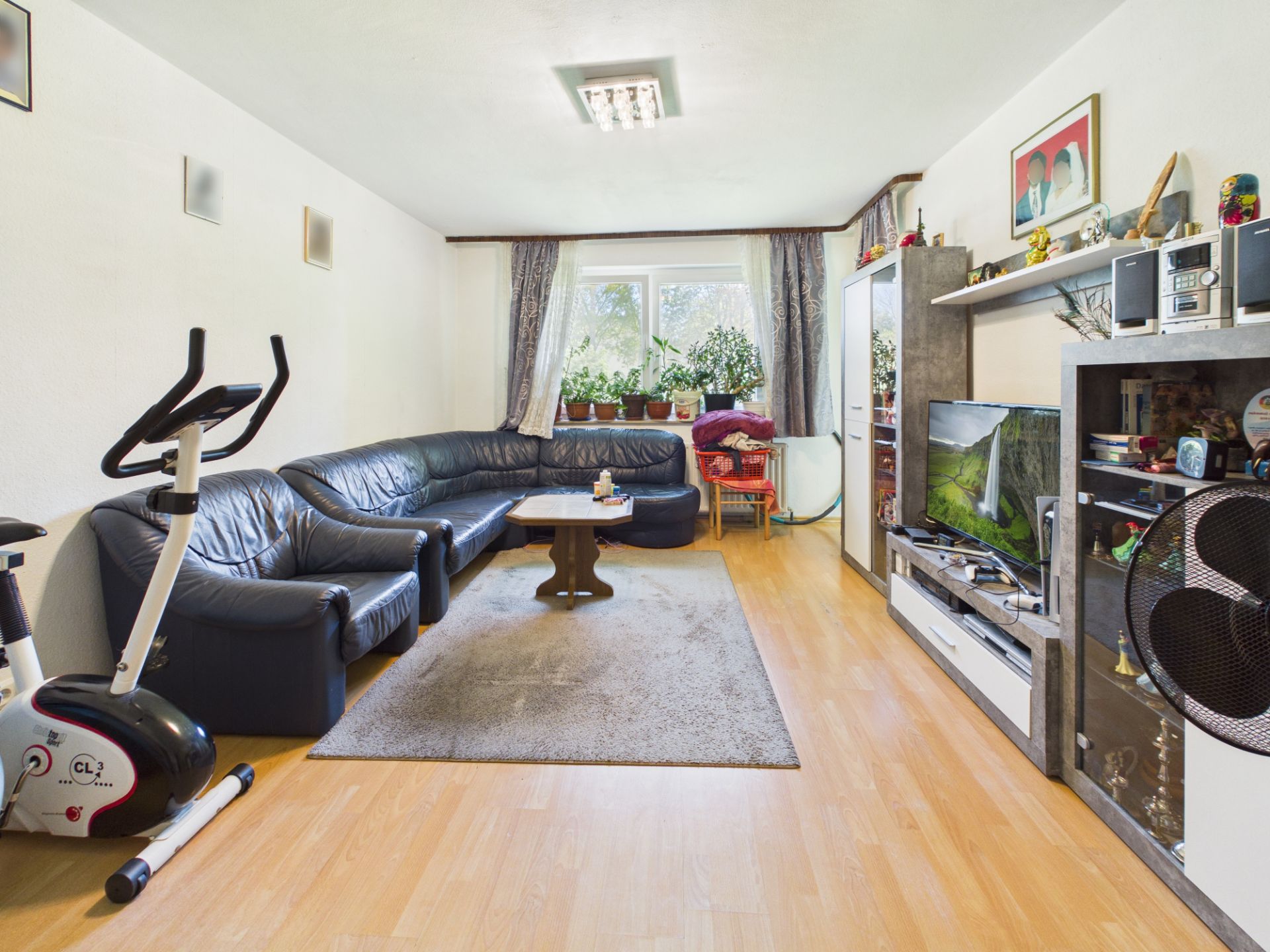 Wohnzimmer mit Ecksofa, Teppich, Fernseher, Heimtrainer und Pflanzen am Fenster.