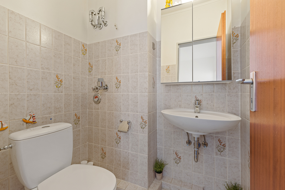 Kleines Badezimmer mit Toilette, Waschbecken, Spiegelschrank und gefliesten Wänden mit Blumenmuster.