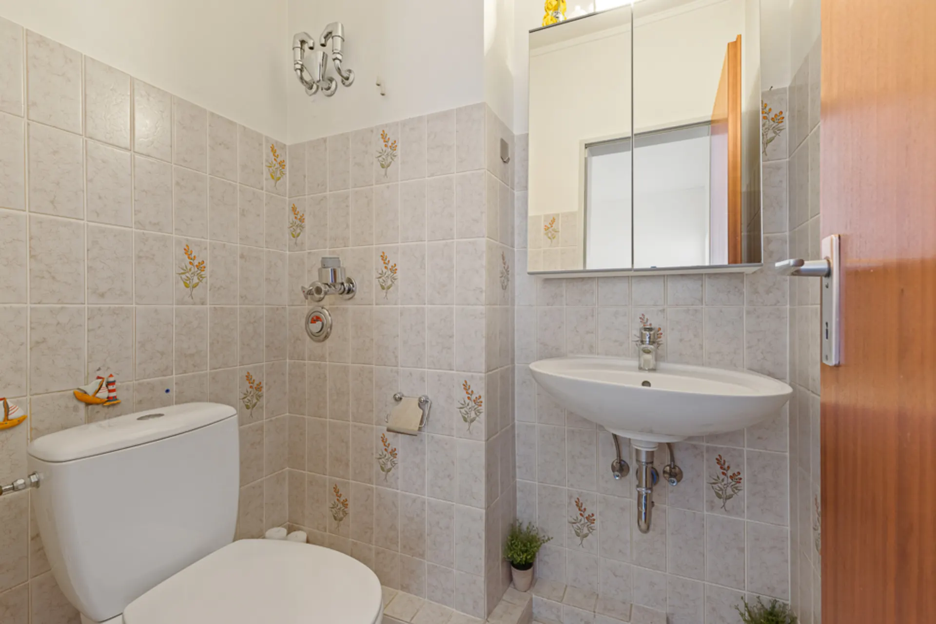 Kleines Badezimmer mit Toilette, Waschbecken, Spiegelschrank und gefliesten Wänden mit Blumenmuster.
