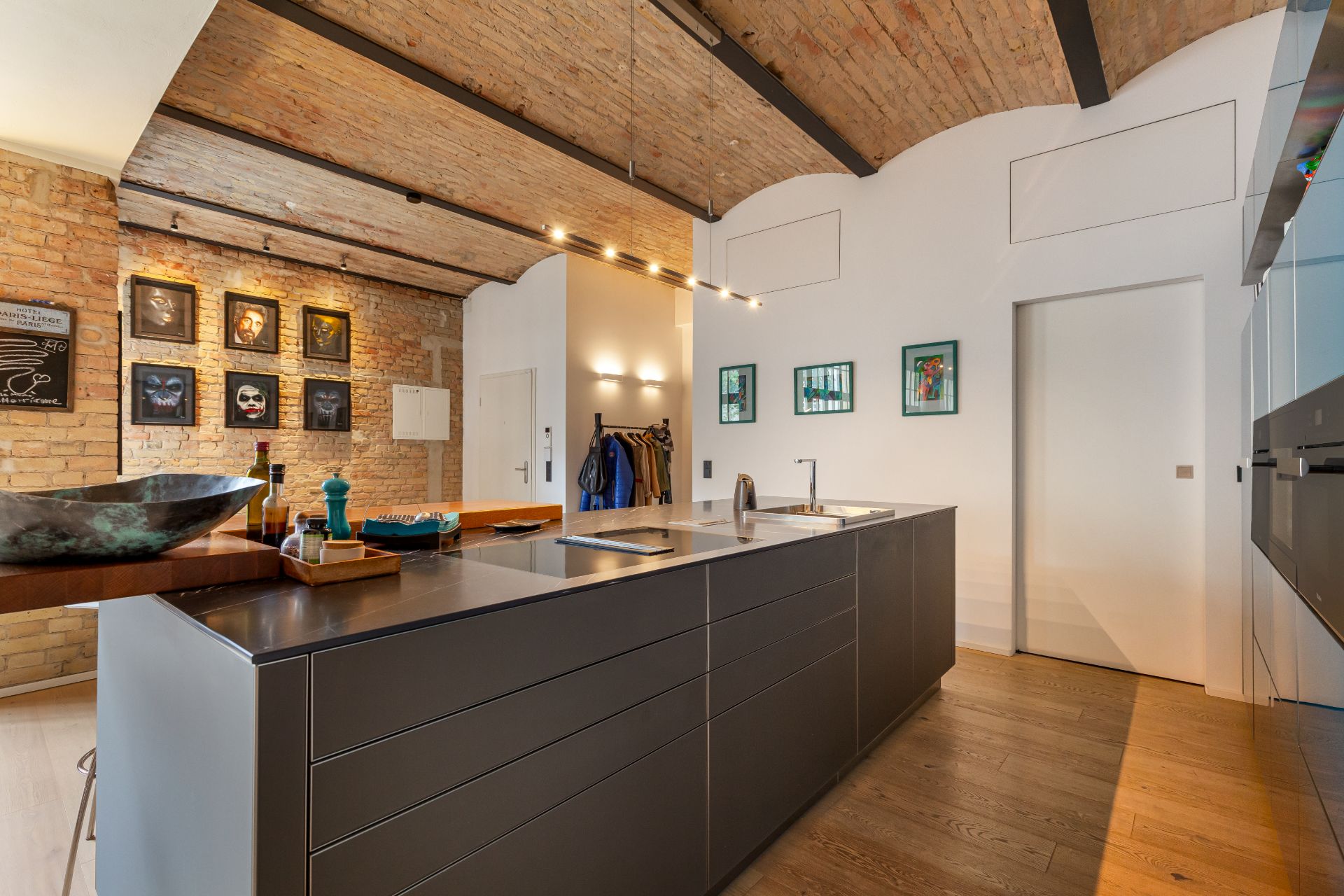 Moderne Küche mit großer Kochinsel, Holzboden und Backsteinwand, einige Bilder an den Wänden.