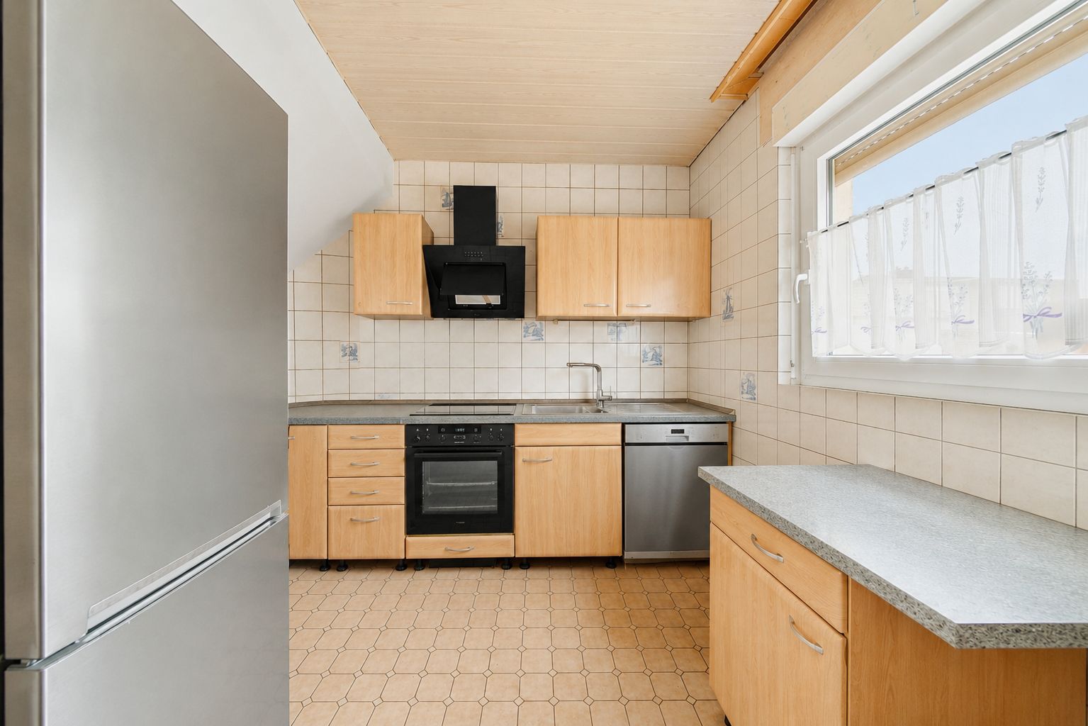 Helle Küche mit Holzfronten, Edelstahlkühlschrank, Herd, Spülmaschine und großem Fenster.