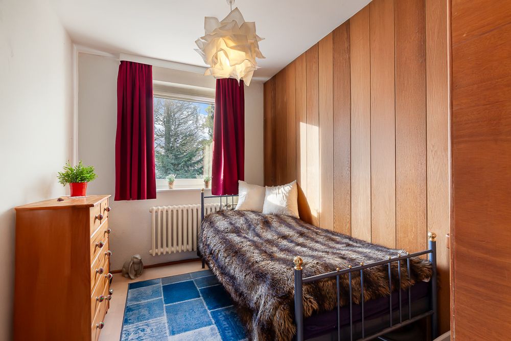 Kleines Schlafzimmer mit Einzelbett, Holzvertäfelung, Kommode und Fenster mit roten Vorhängen.