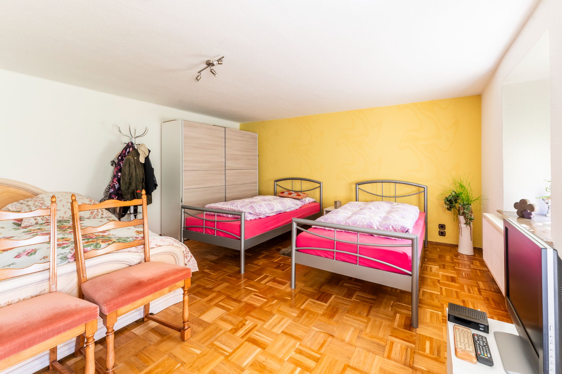 Helles Schlafzimmer mit zwei Einzelbetten, einem Doppelbett, Schrank und Fernseher auf Parkettboden.