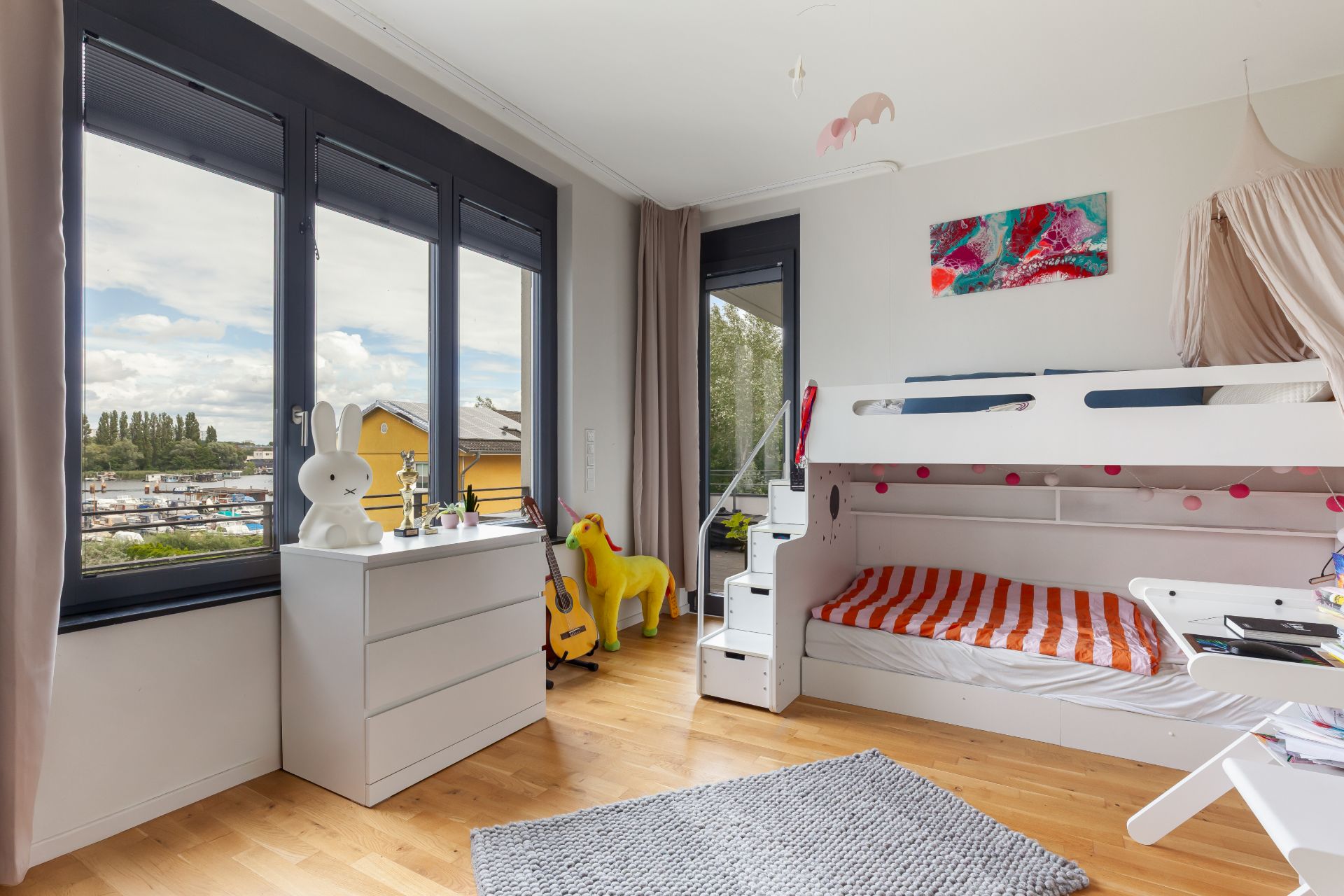 Helles Kinderzimmer mit Etagenbett, Spielzeug, Kommode und großem Fenster.