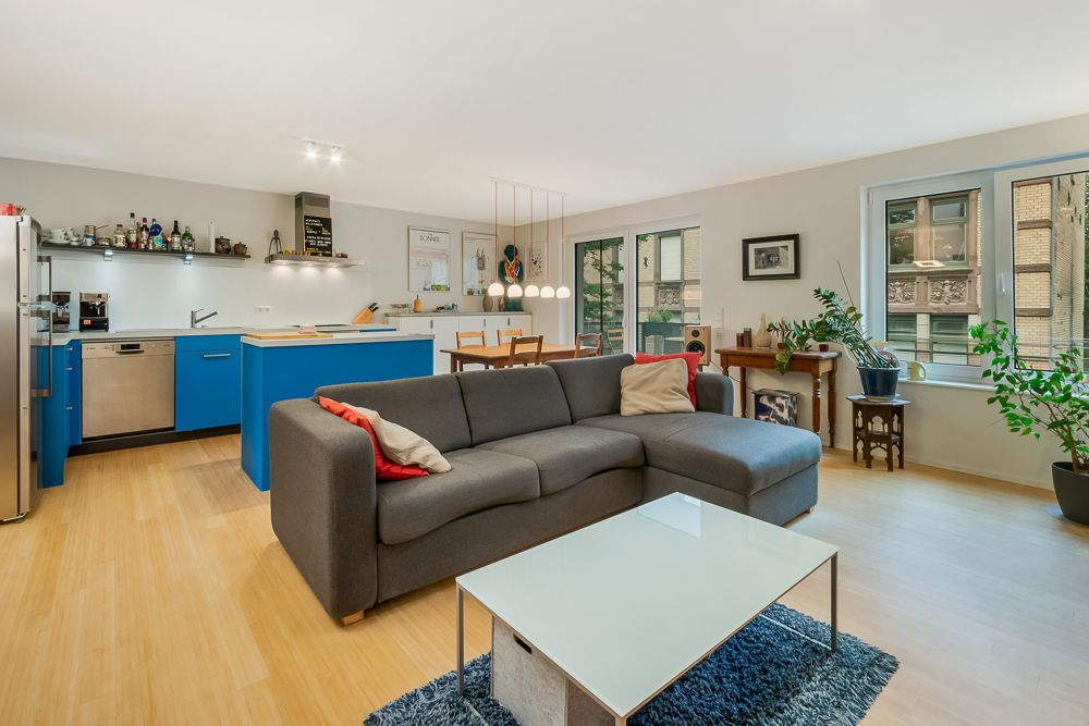 Modernes Wohnzimmer mit grauem Sofa, blauem Küchenbereich und großen Fenstern, Holzboden, Pflanzen.