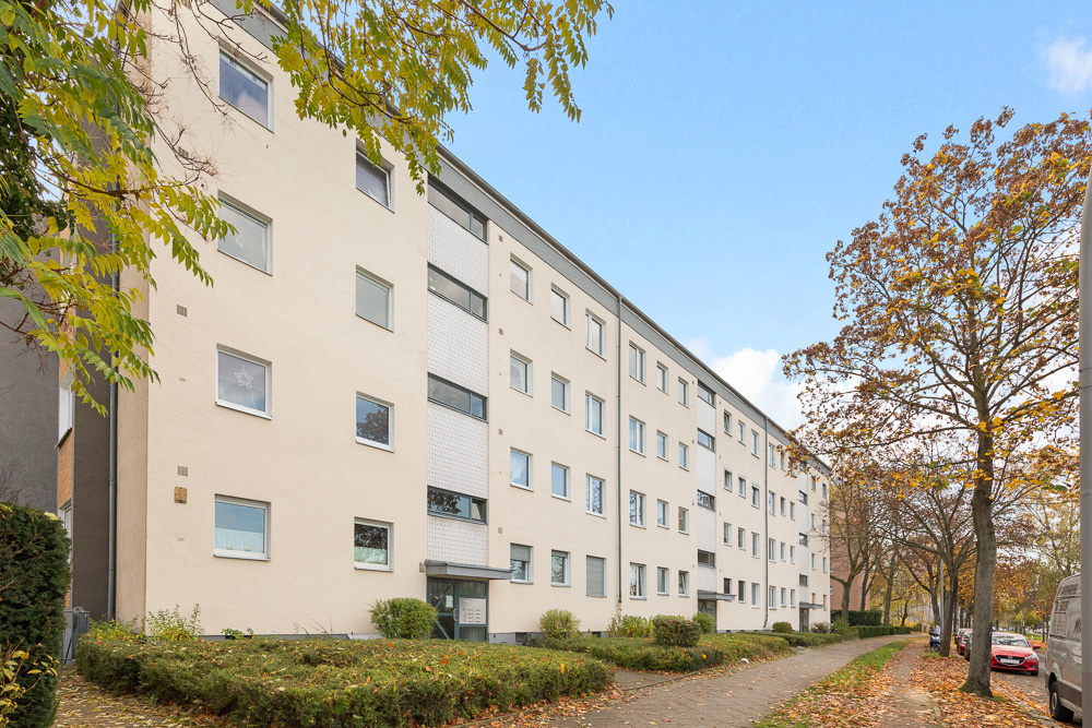 Mehrstöckiges Wohnhaus mit hellen Fassaden, Gehweg und Bäumen im Vordergrund.