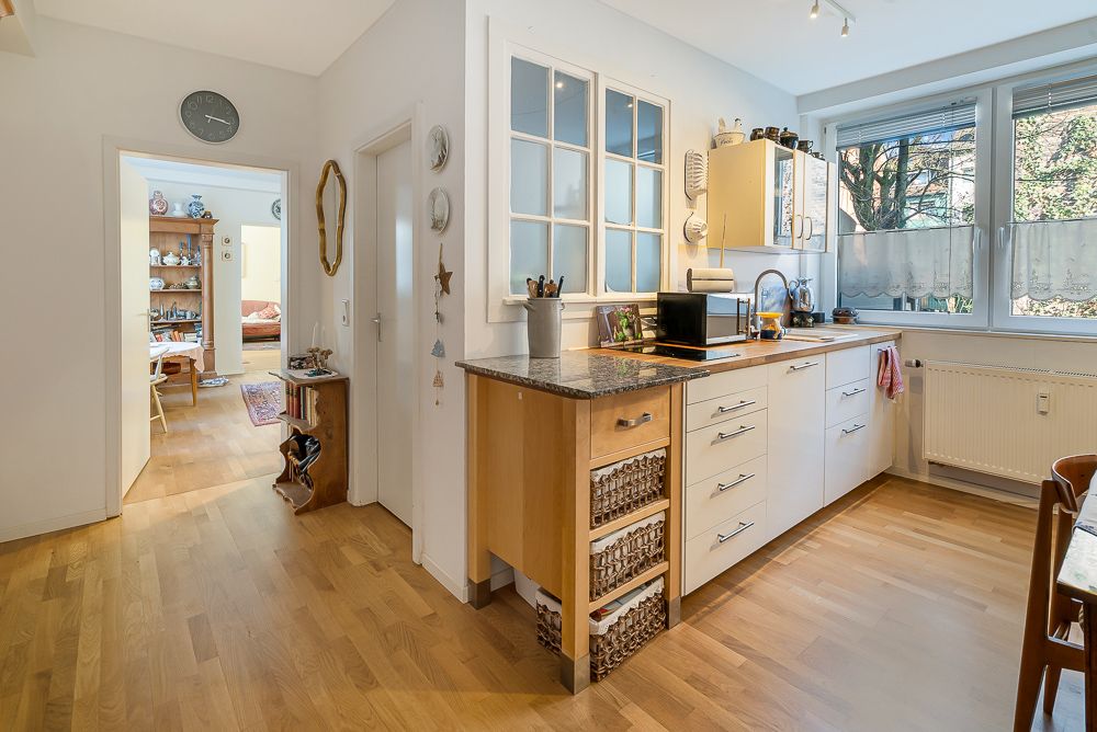 Helle Küche mit Holzfußboden, weißen Schränken und großem Fenster, Blick in ein weiteres Zimmer.