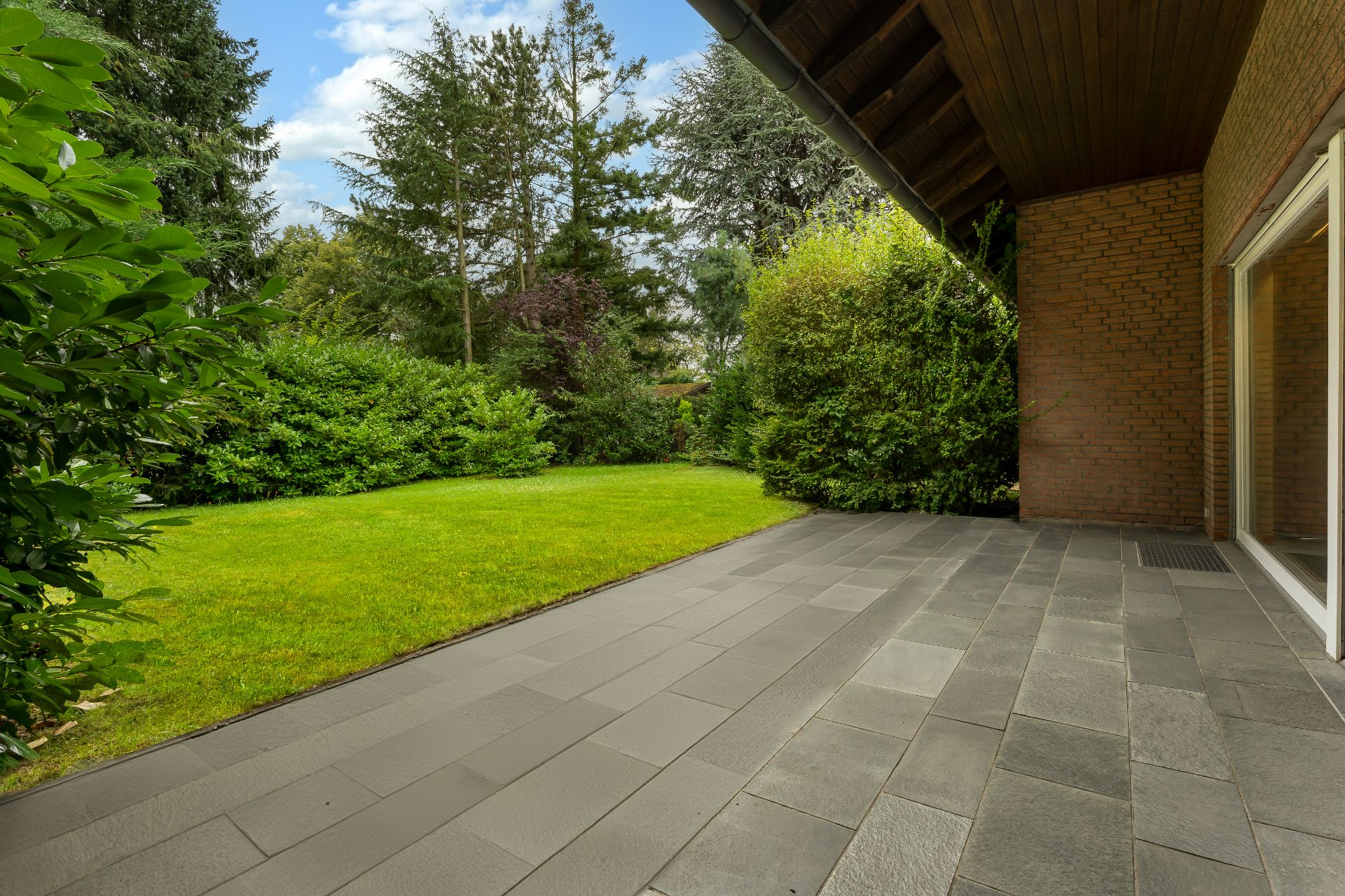 Überdachte Terrasse mit grauen Fliesen und Blick auf einen grünen Garten mit Büschen und Bäumen.