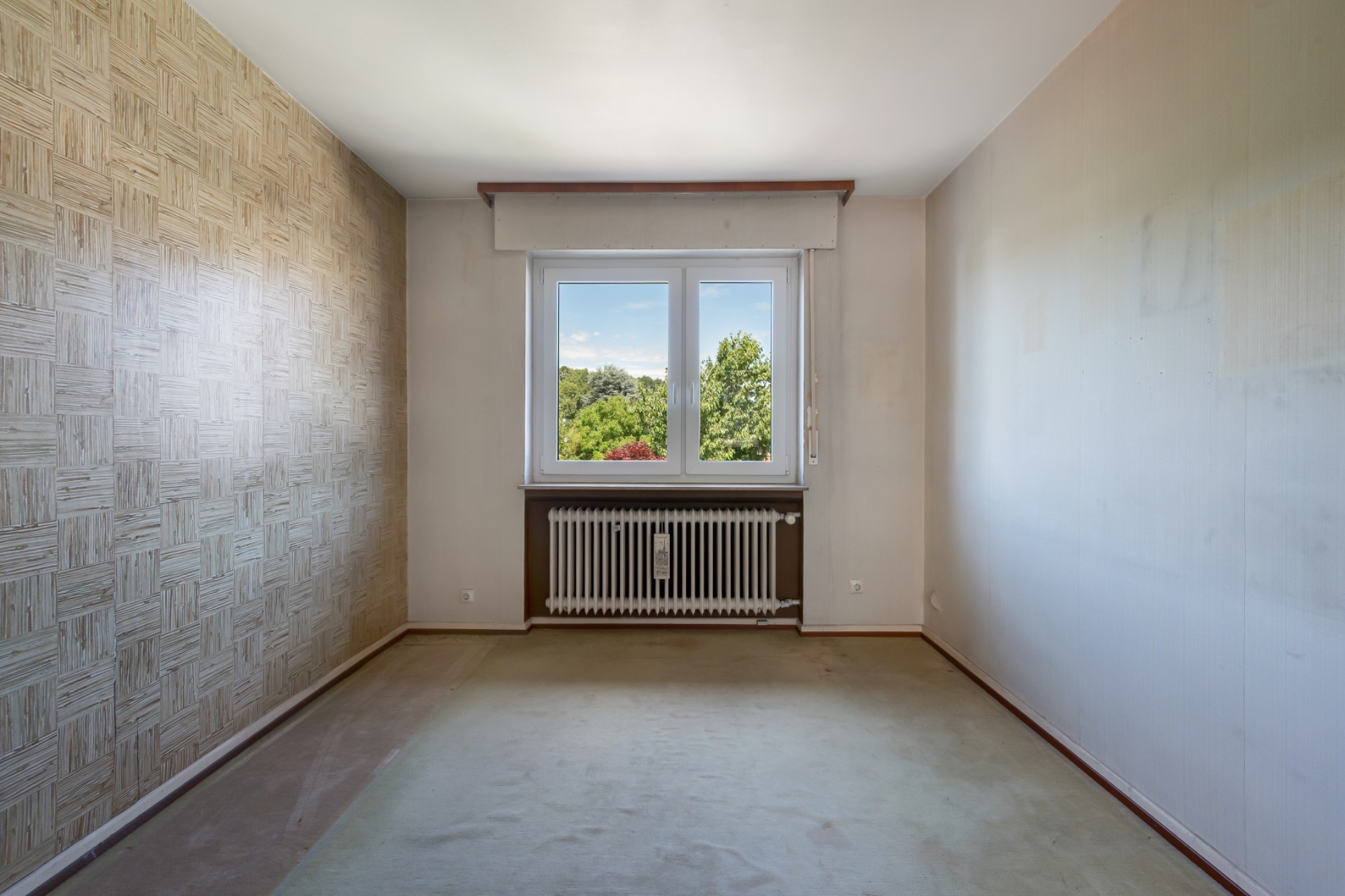 Leeres Zimmer mit Teppichboden, einem Fenster und Heizung unter dem Fenster.