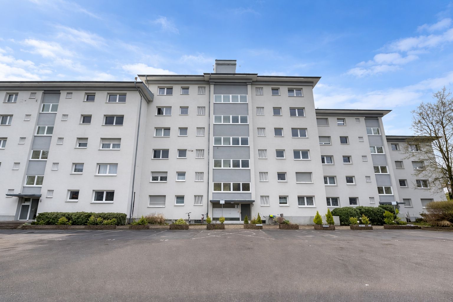 Mehrstöckiges Wohnhaus mit vielen Fenstern und kleinem Vorplatz, davor Parkfläche.