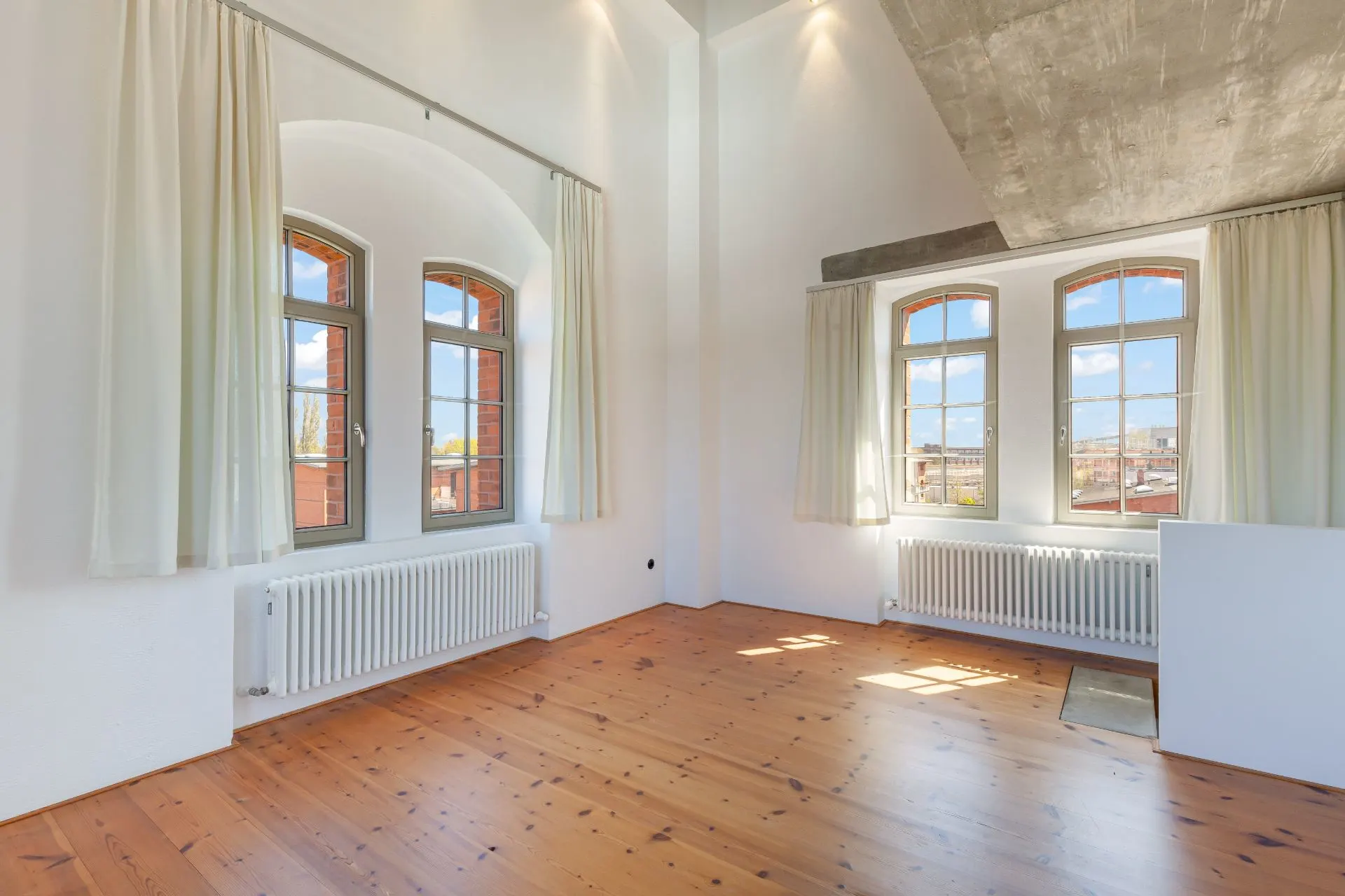 Helles Zimmer mit Holzboden, großen Fenstern, weißen Vorhängen und Heizkörpern unter den Fenstern.