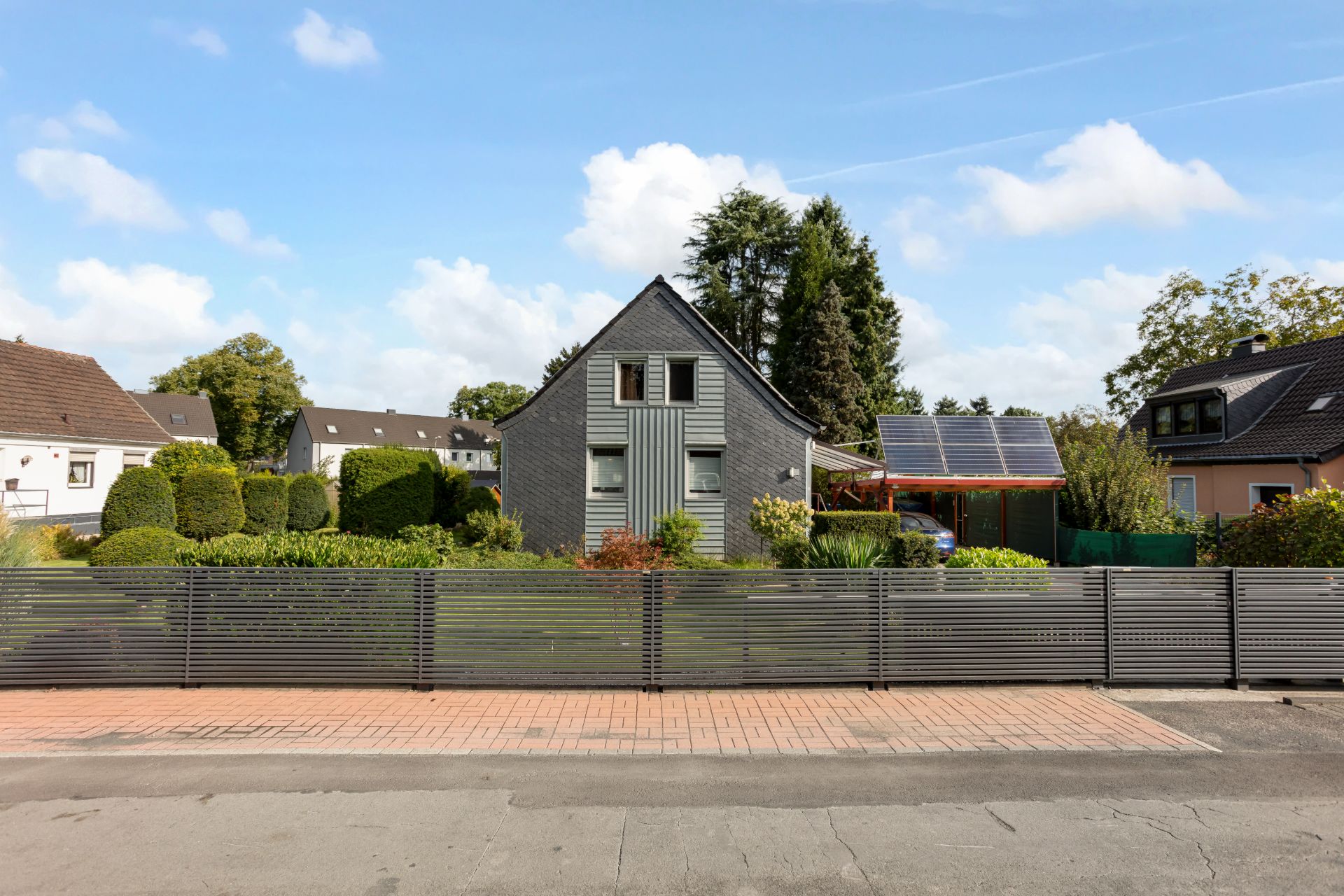 Modernes Einfamilienhaus mit spitzem Dach, Garten und Metallzaun im Vordergrund.