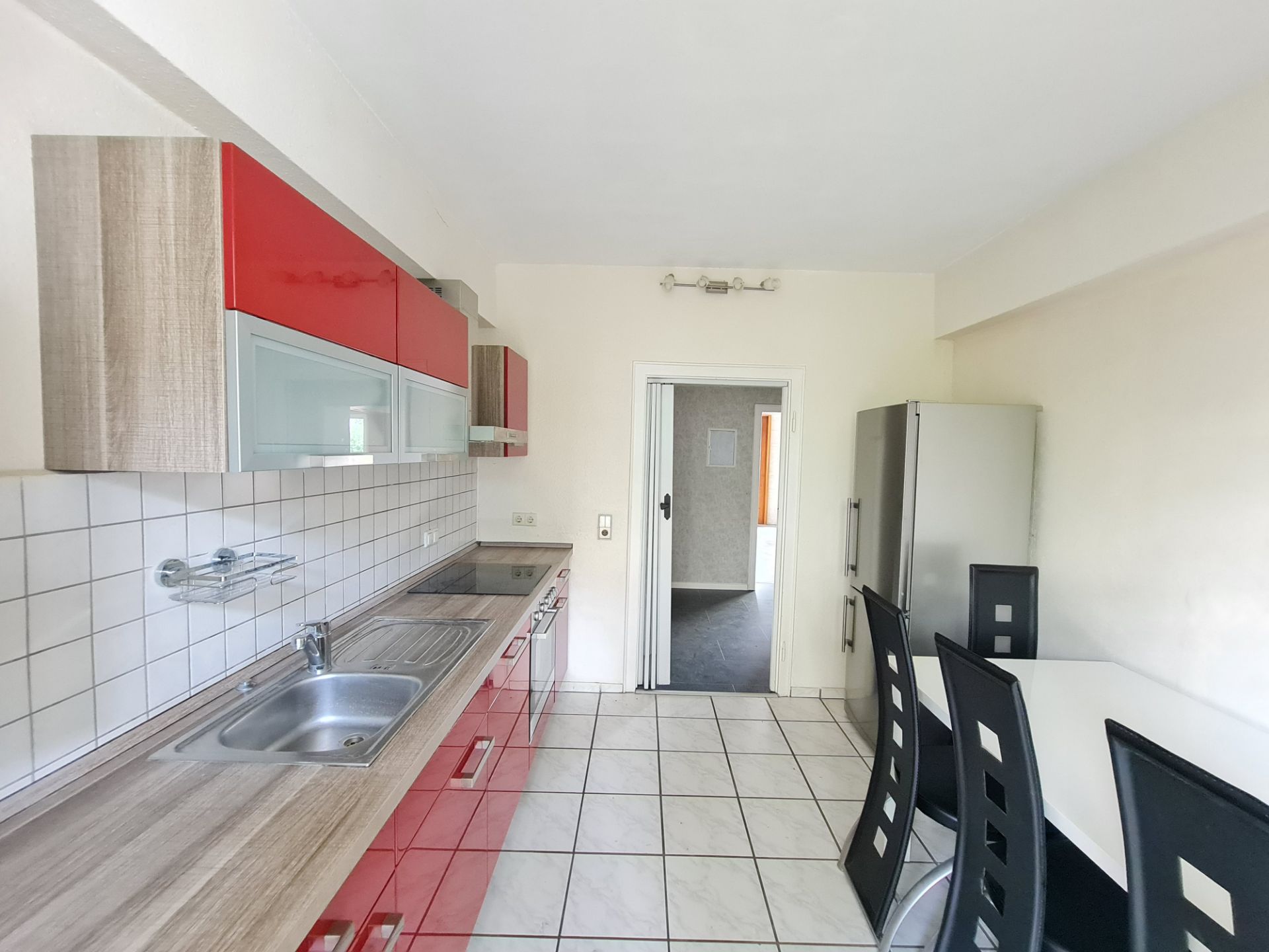 Moderne Küche mit roten Schränken, Esstisch und vier schwarzen Stühlen, Fliesenboden, Kühlschrank.