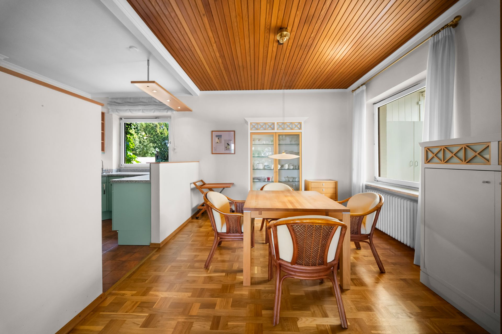 Esszimmer mit Holztisch, fünf Stühlen, Vitrine und Holzdecke, großes Fenster an der rechten Wand.
