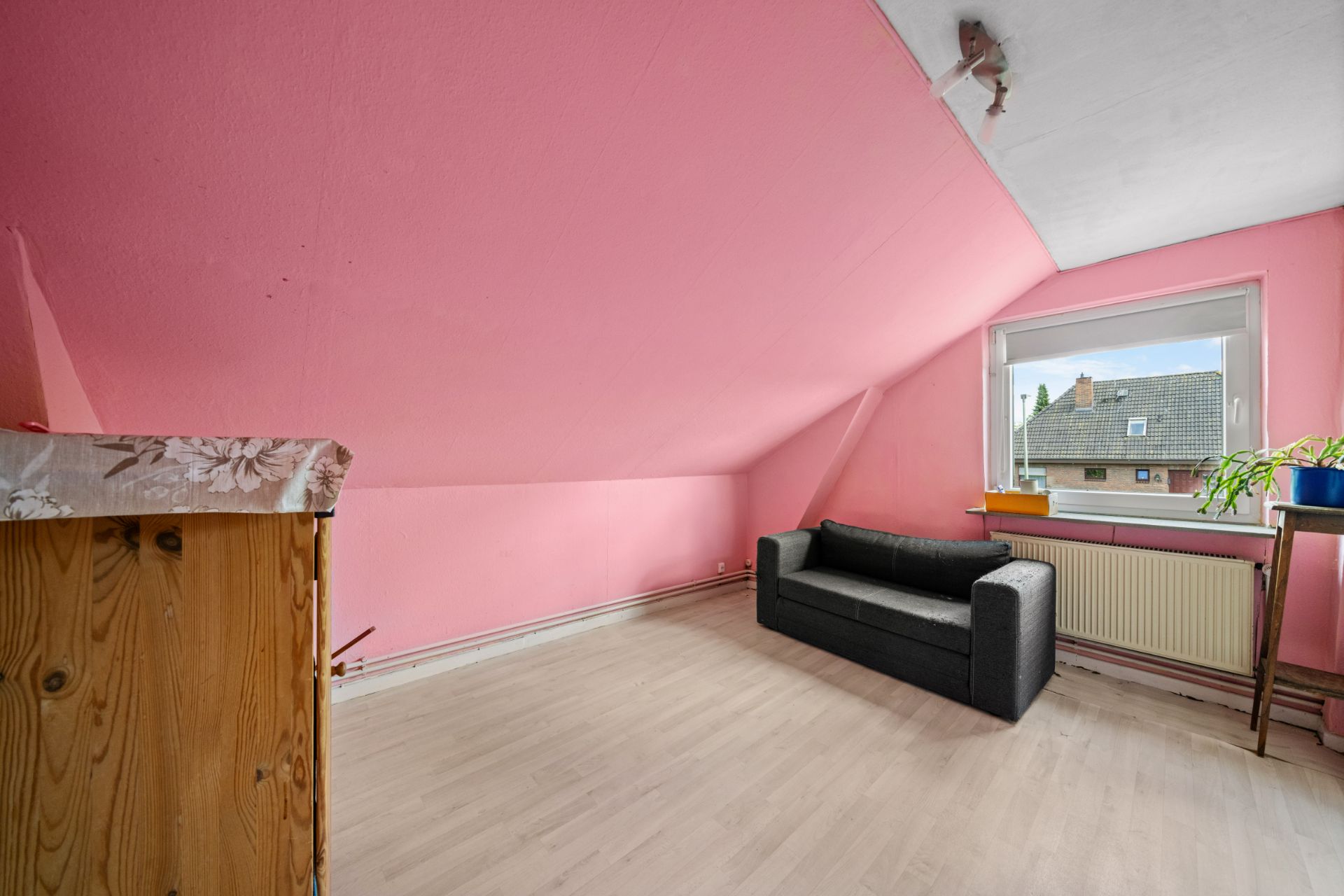 Zimmer mit pinker Dachschräge, grauem Sofa, Holzkommode und Fenster mit Blick nach draußen.
