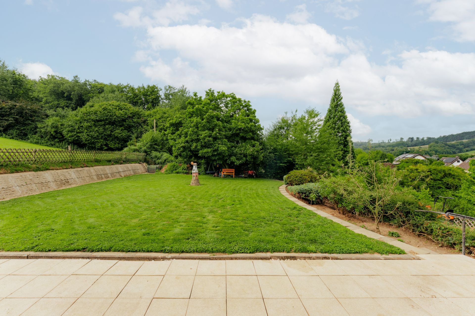 Großer Garten mit Rasenfläche, Bäumen, einer Bank und einer Statue, angrenzend an eine Terrasse.