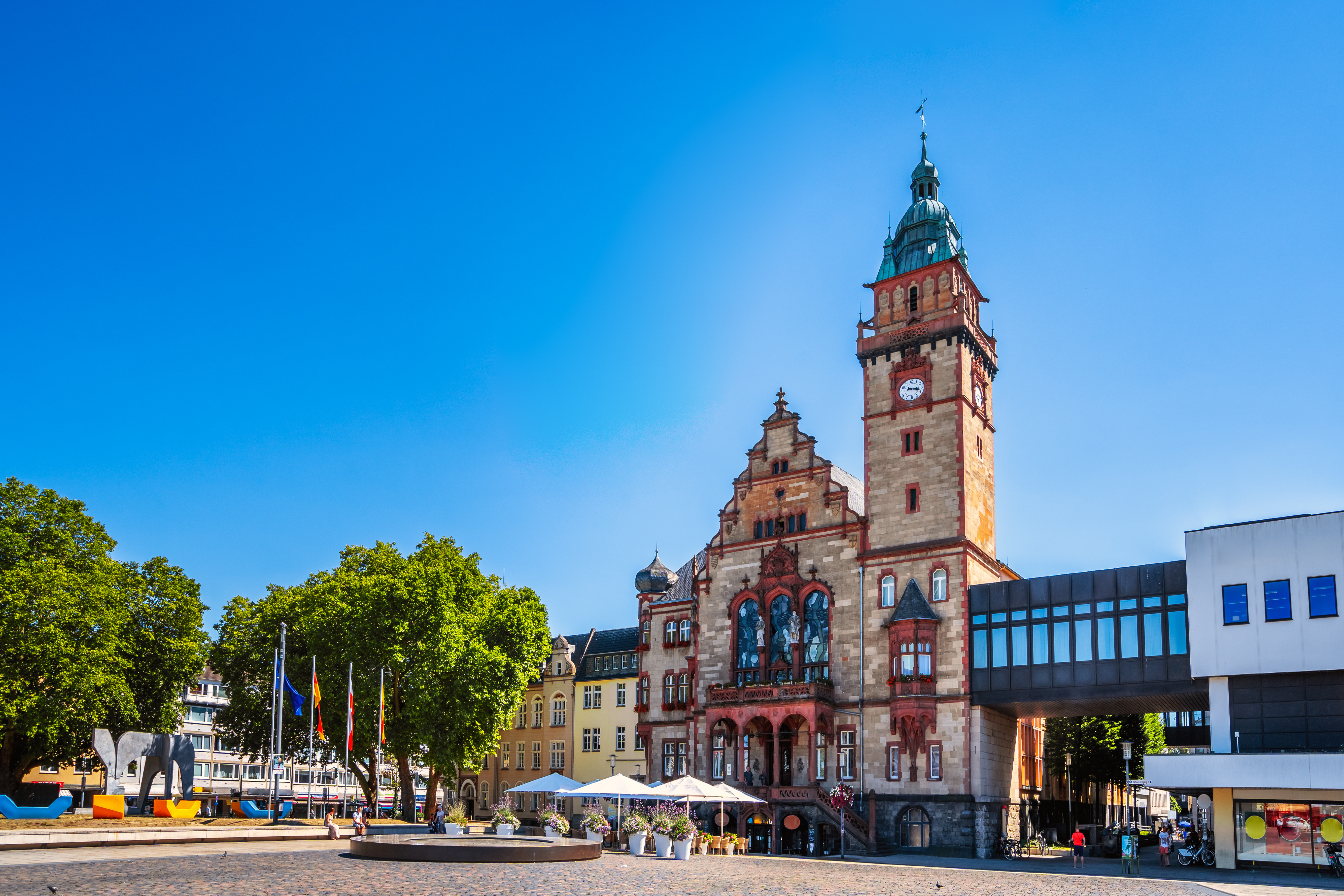 Historisches Gebäude mit Uhrturm auf einem sonnigen Platz mit Bäumen und Fahnen.