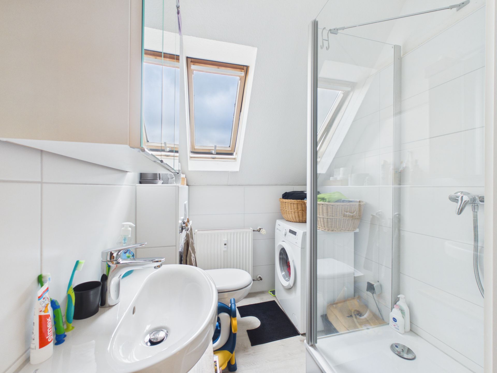 Helles Badezimmer mit Waschbecken, Dusche, Toilette, Waschmaschine und zwei Dachfenstern.