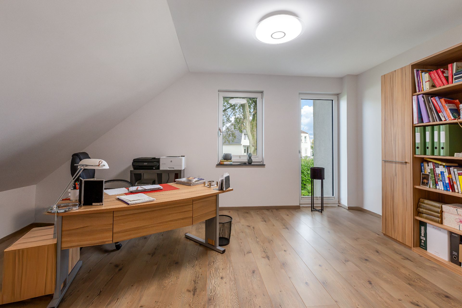 Helles Arbeitszimmer mit Schreibtisch, Regal, Holzfußboden und zwei Fenstern.