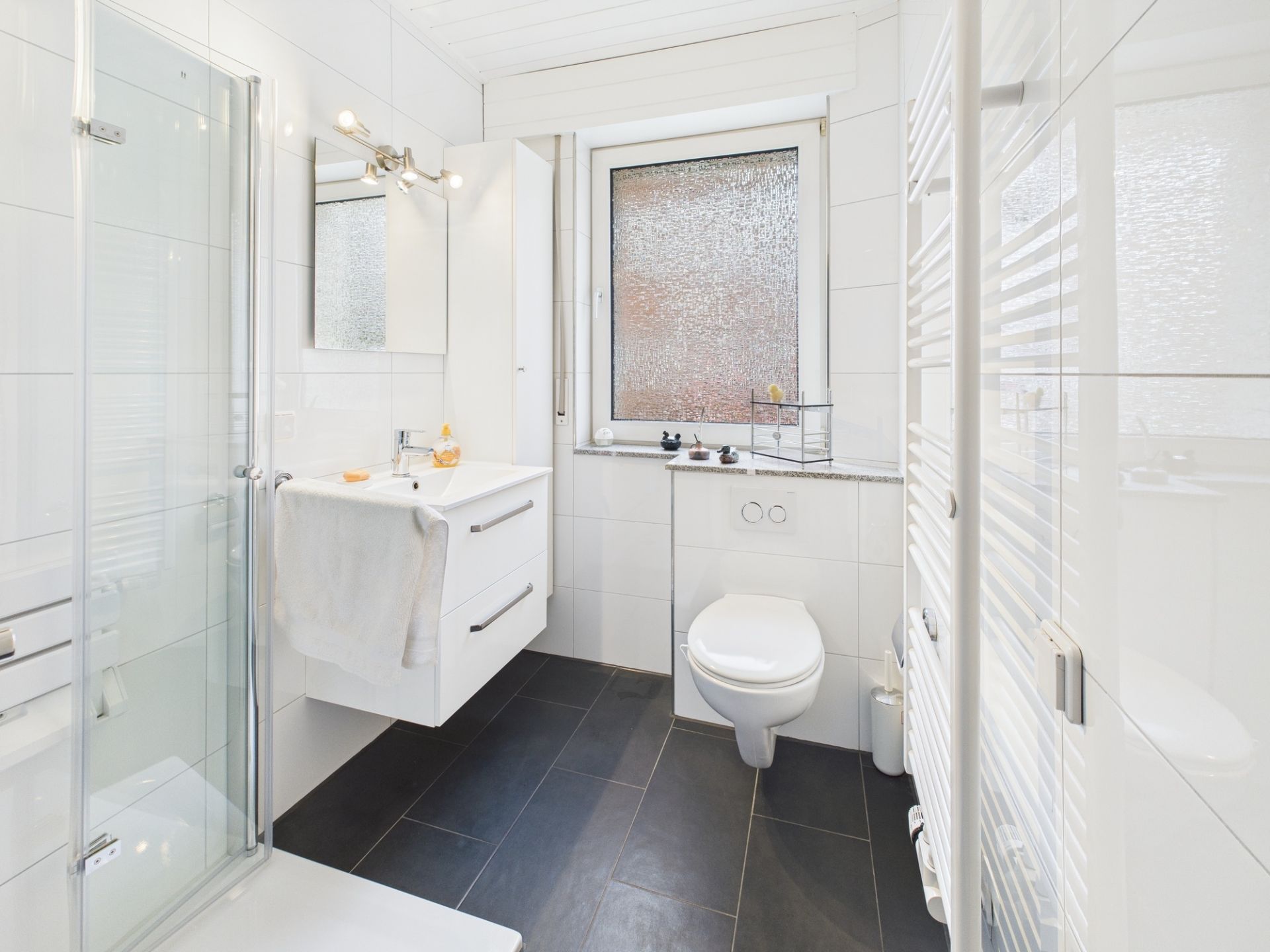 Modernes, weiß gefliestes Badezimmer mit Dusche, Waschbecken, WC und Fenster.