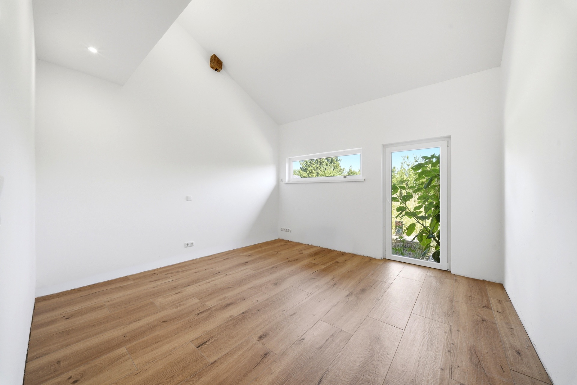 Heller, leerer Raum mit Holzfußboden, schräger Decke, Fenster und Glastür zu grünem Garten.