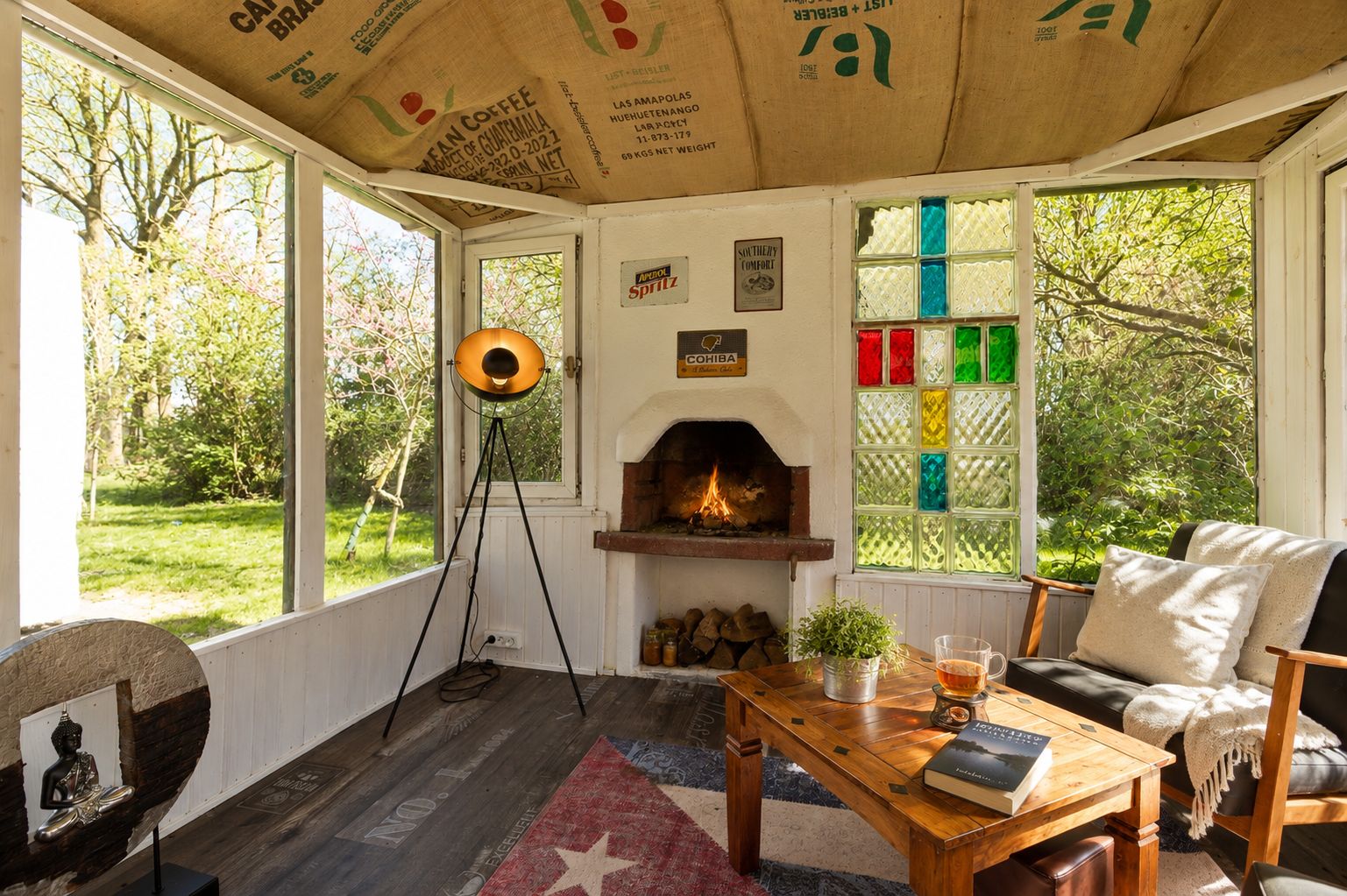 Gemütlicher Wohnraum mit Kamin, Holztisch, Sessel und buntem Fenster, umgeben von Garten.