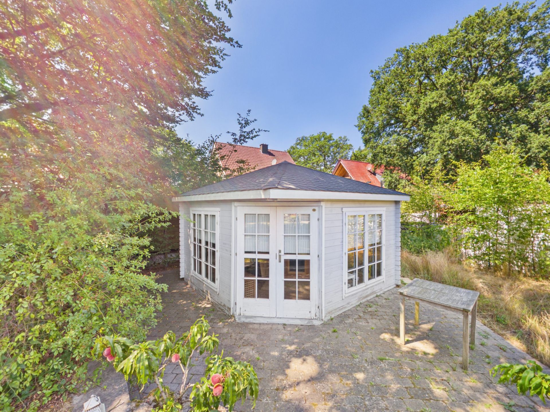 Kleines weißes Gartenhaus mit großen Fenstern und Doppeltür auf einer Terrasse, umgeben von Bäumen und Pflanzen.