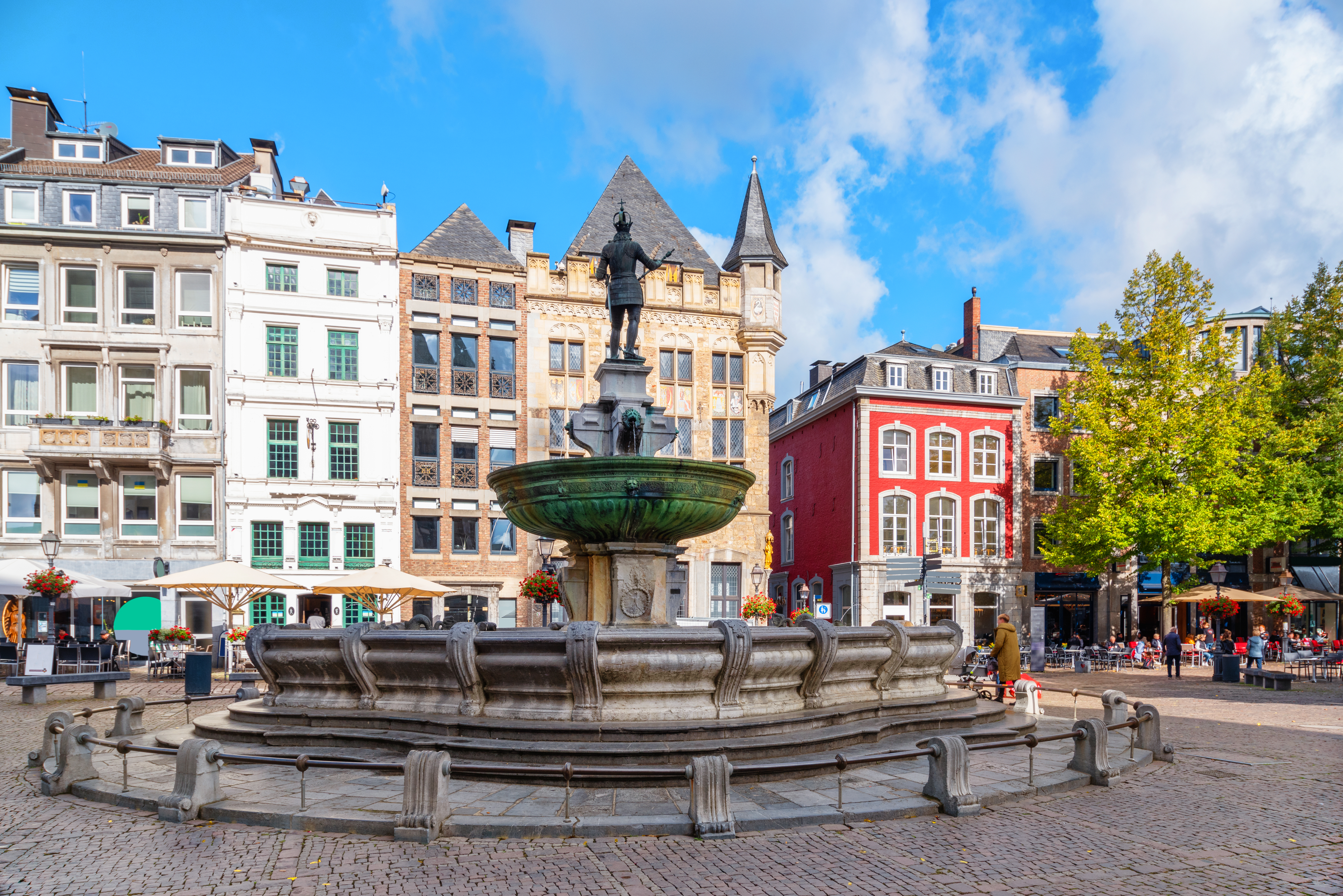 Großer Brunnen auf einem Platz mit historischen Gebäuden und Cafés im Hintergrund.