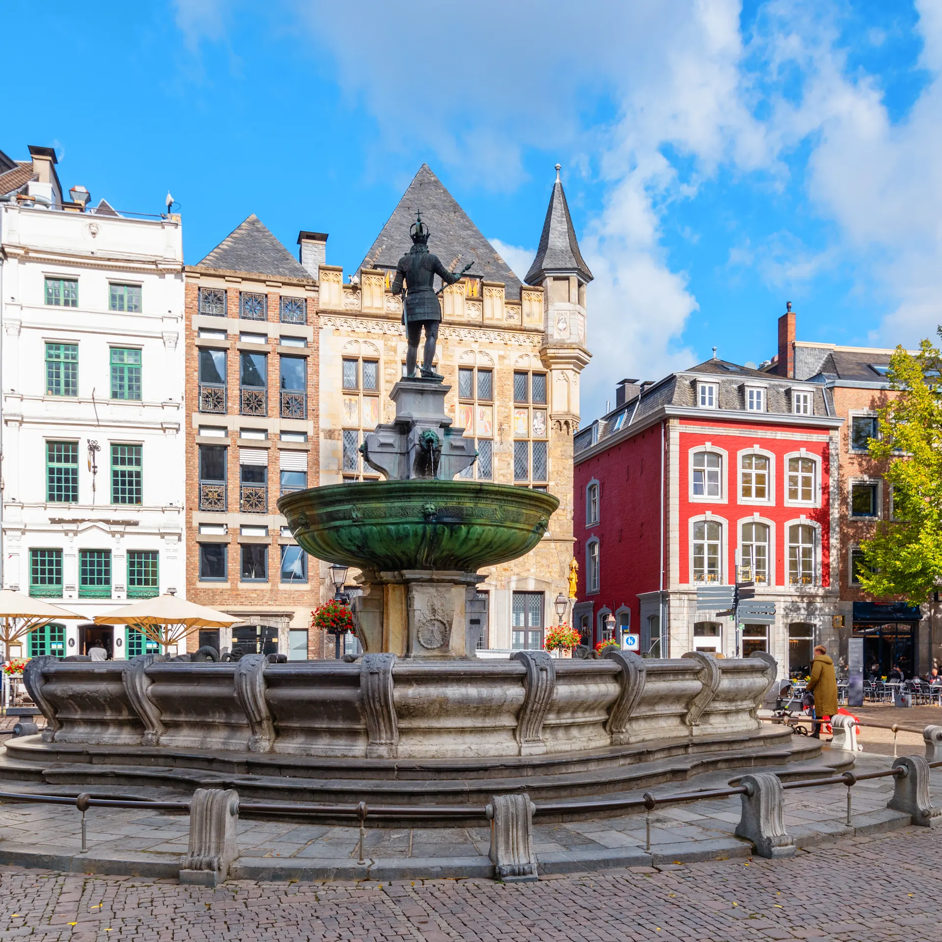 Großer Brunnen auf einem Platz mit historischen Gebäuden und Cafés im Hintergrund.