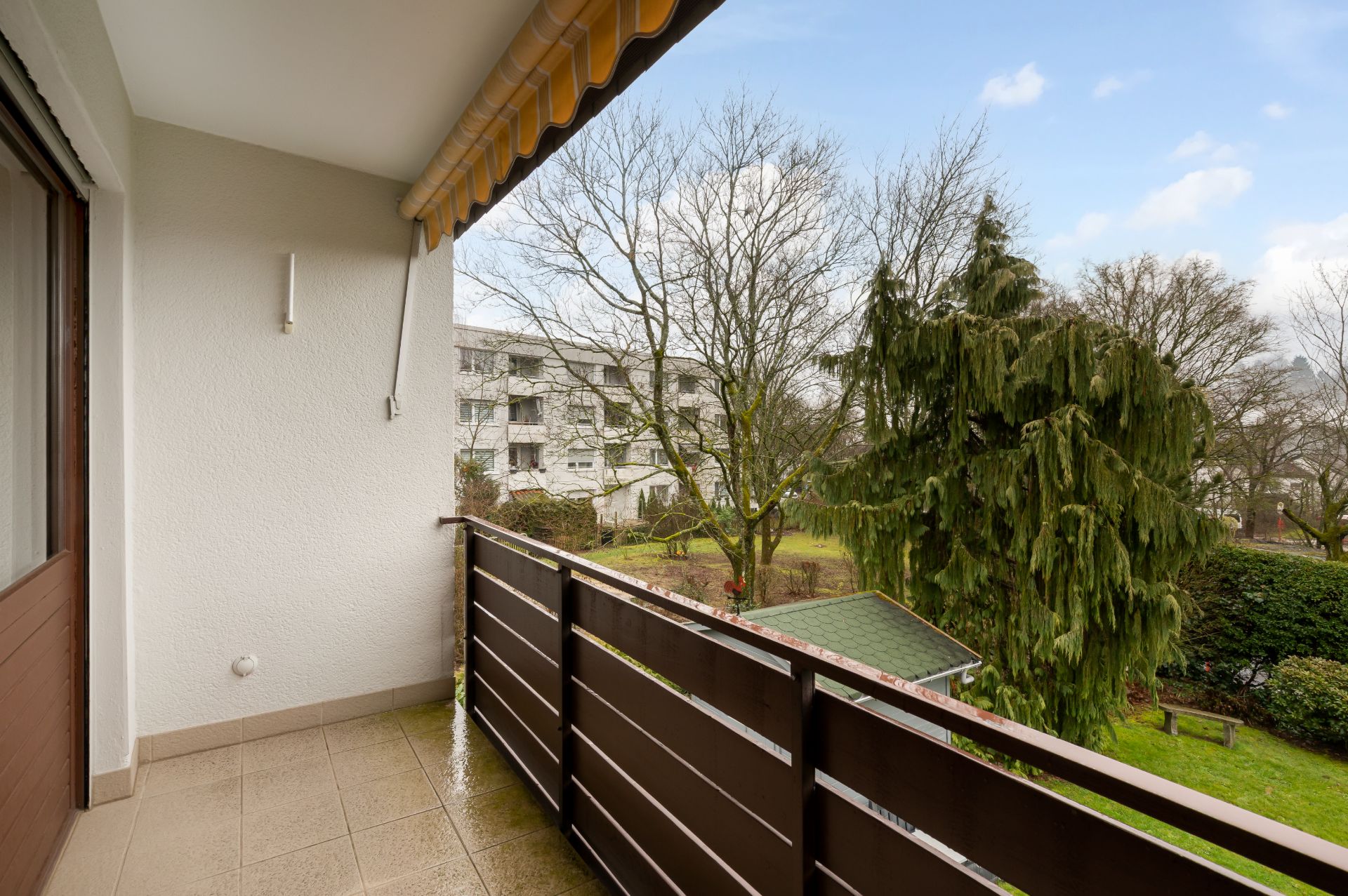 Überdachter Balkon mit braunem Geländer, Blick auf Bäume und benachbarte Wohngebäude.