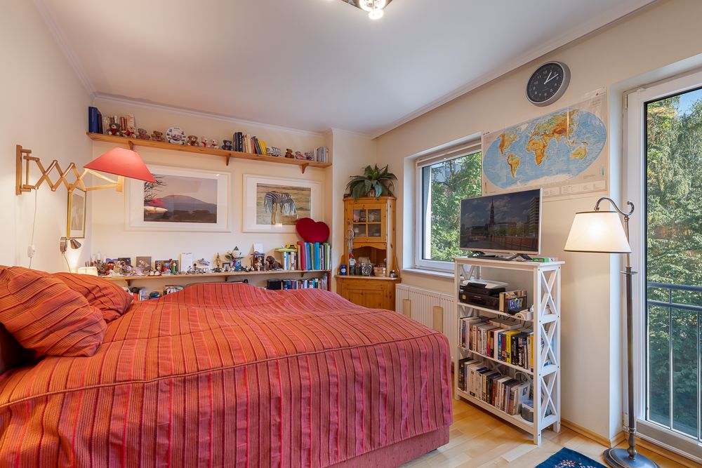 Gemütliches Schlafzimmer mit rotem Bett, Regal, Büchern, Fernseher und großem Fenster.