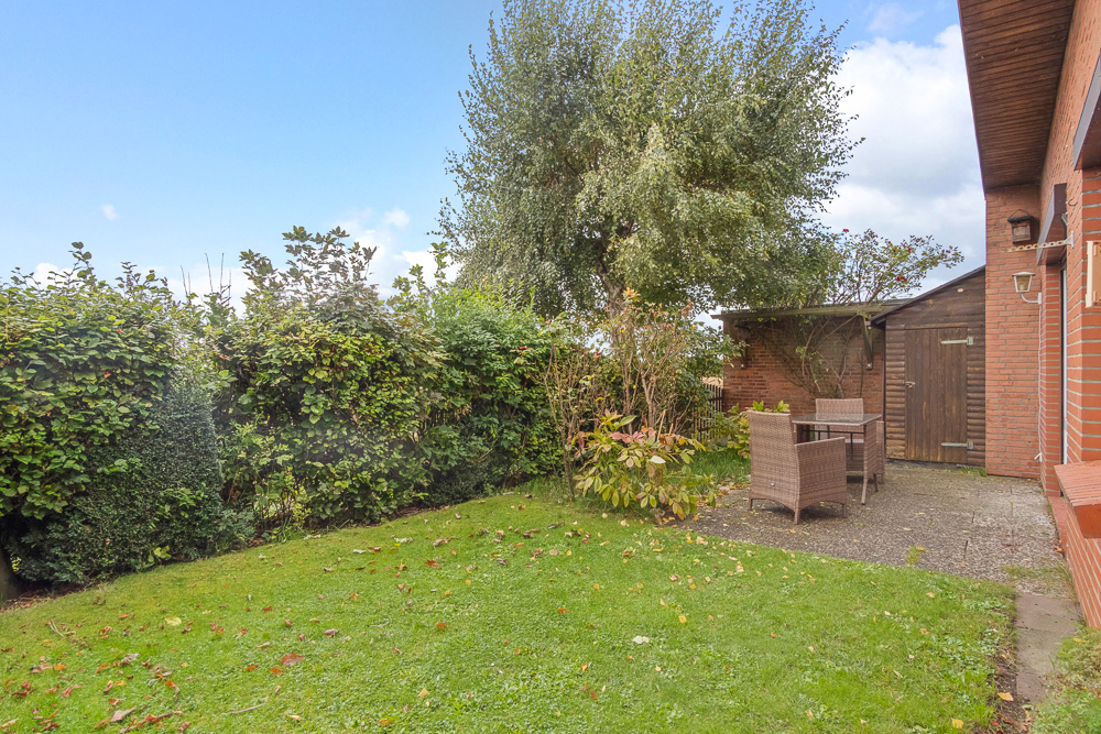 Garten mit Rasen, Terrasse, Gartentisch und Stühlen neben einem Ziegelhaus und einer Holzhütte.