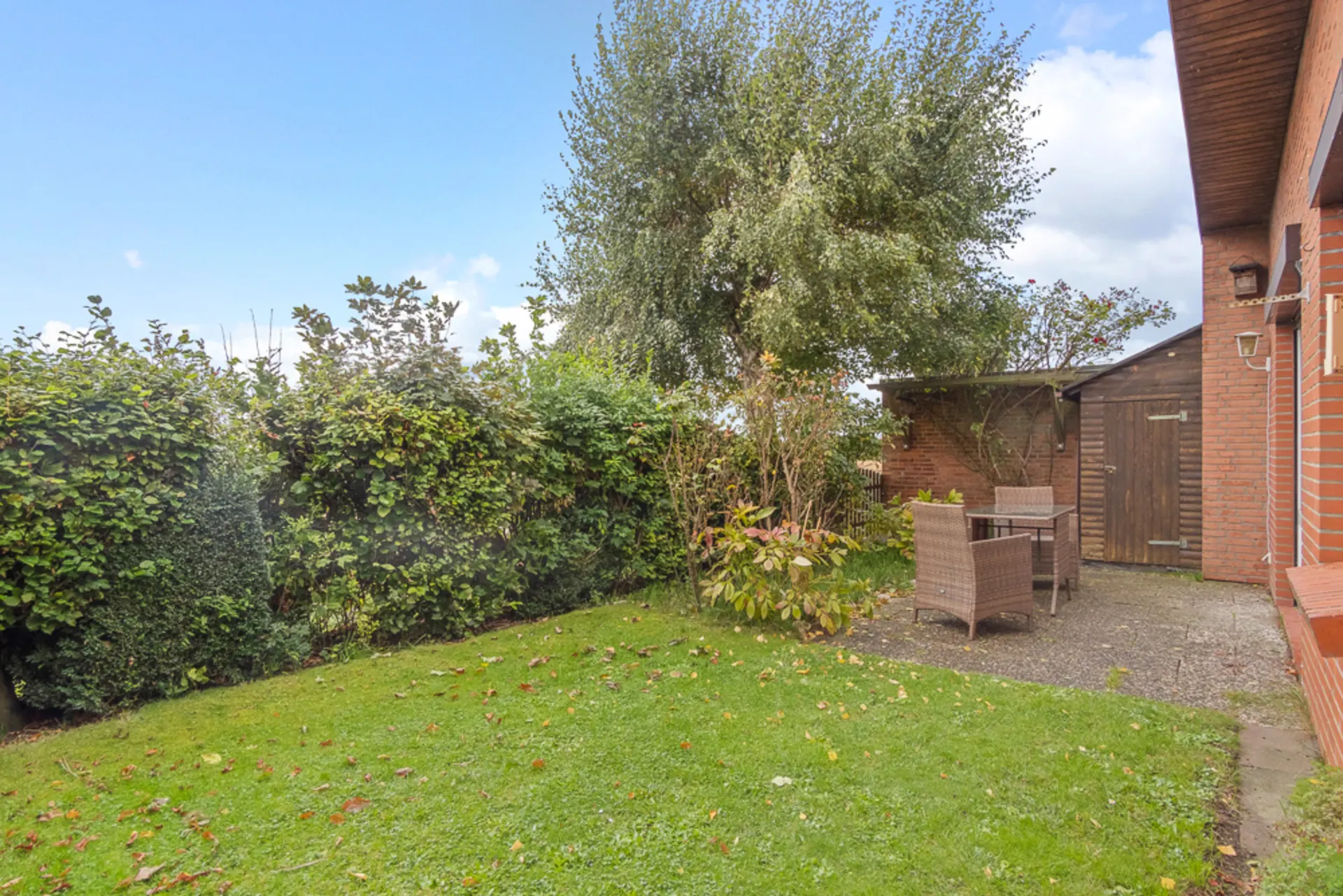 Garten mit Rasen, Terrasse, Gartentisch und Stühlen neben einem Ziegelhaus und einer Holzhütte.