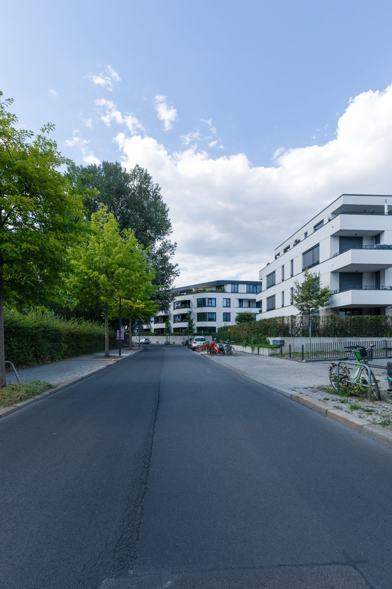 Moderne Wohngebäude an einer ruhigen Straße mit geparkten Fahrrädern und Autos, umgeben von Bäumen.