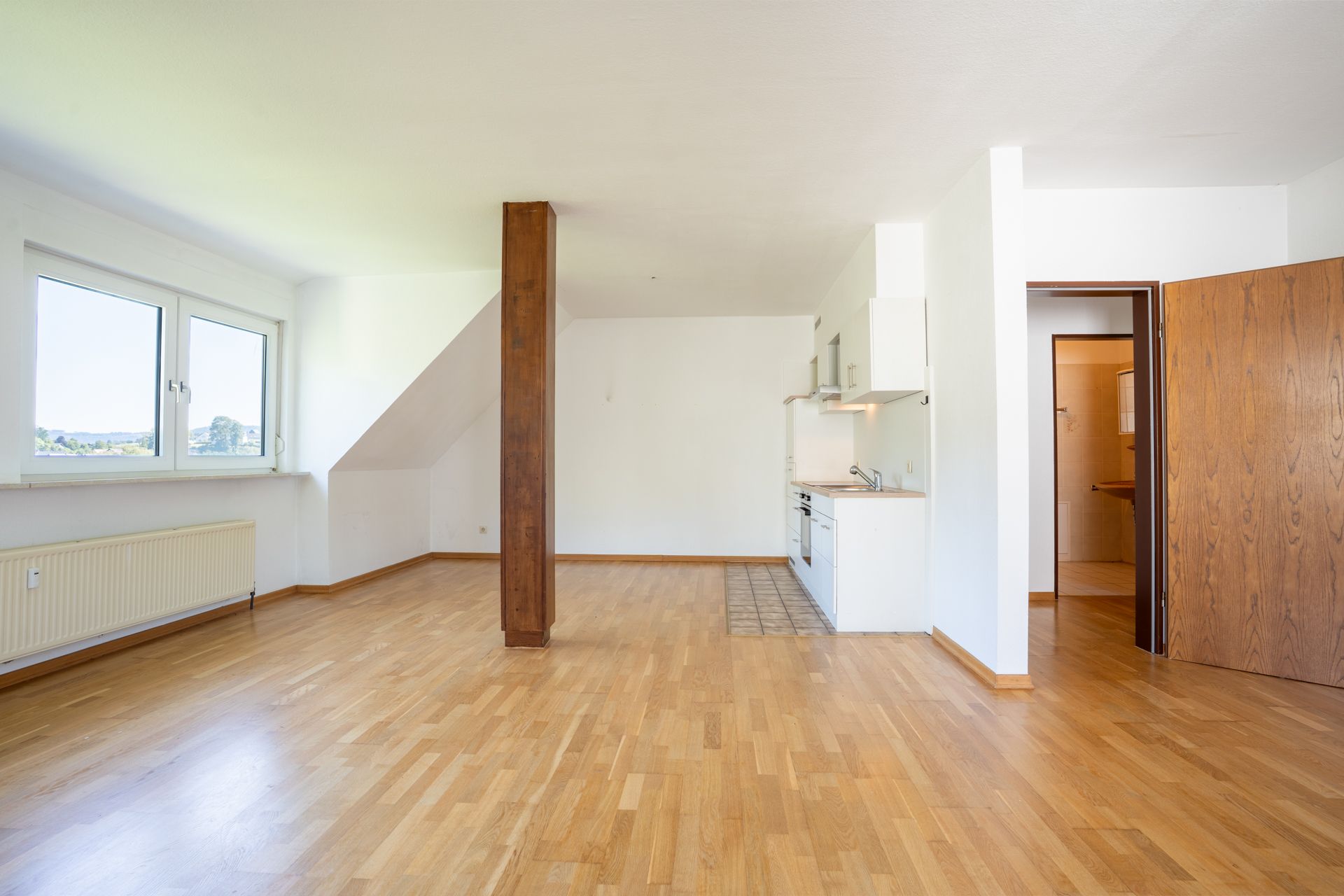 Heller Raum mit Parkettboden, offenem Küchenbereich, Fenstern und Holzsäule.