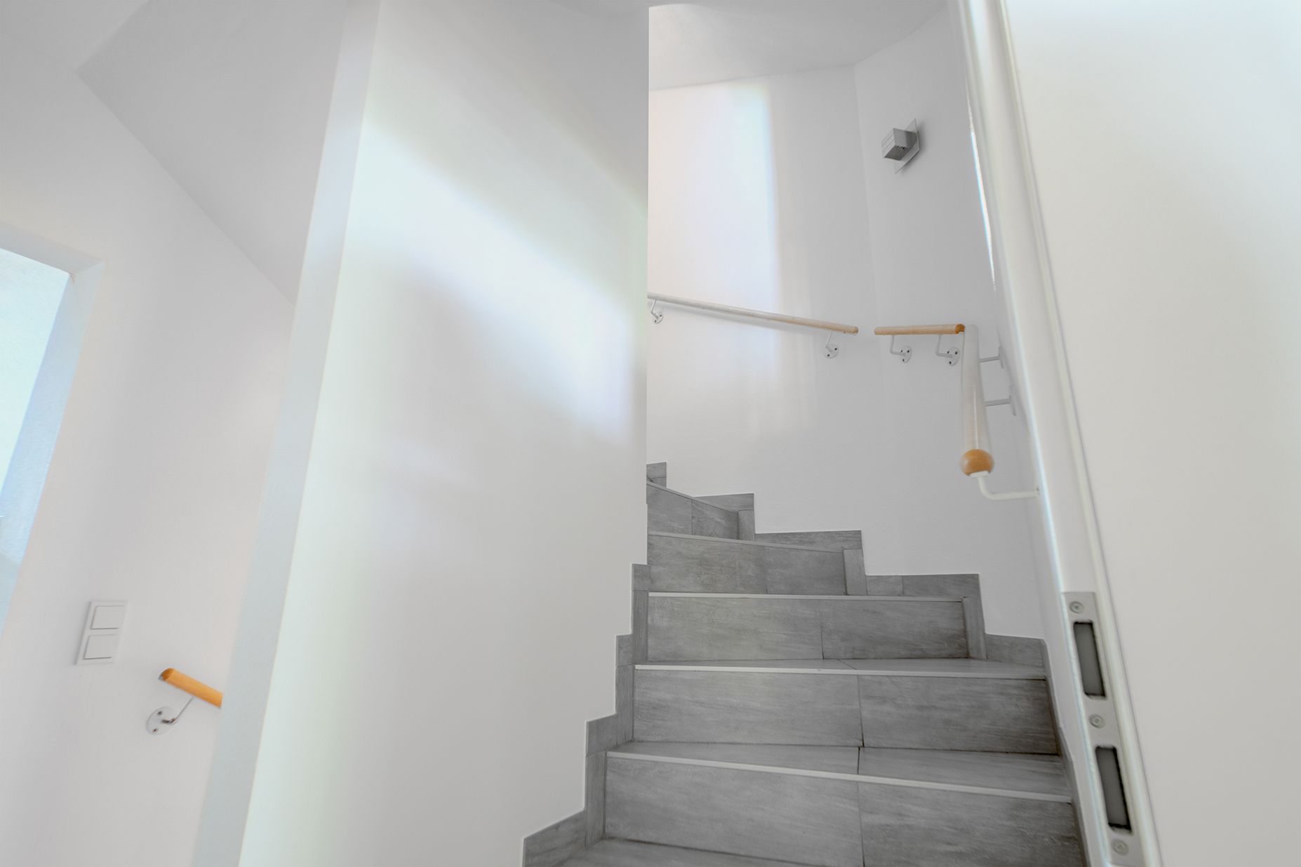 Graue Treppe mit Holzhandläufen in einem hellen, weißen Treppenhaus.