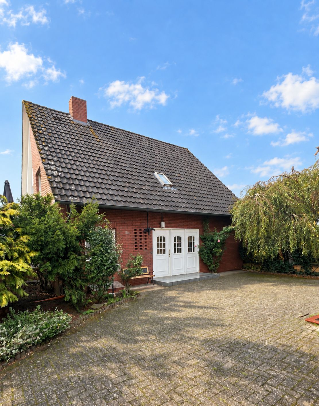 Backsteinwohnhaus mit dunklem Satteldach, weißen Türen und gepflastertem Vorplatz, umgeben von Büschen.