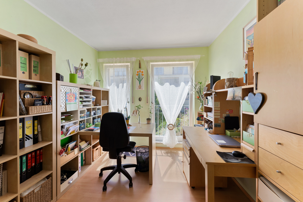 Helles Heimbüro mit zwei Schreibtischen, Regalen und großen Fenstern mit Gardinen.