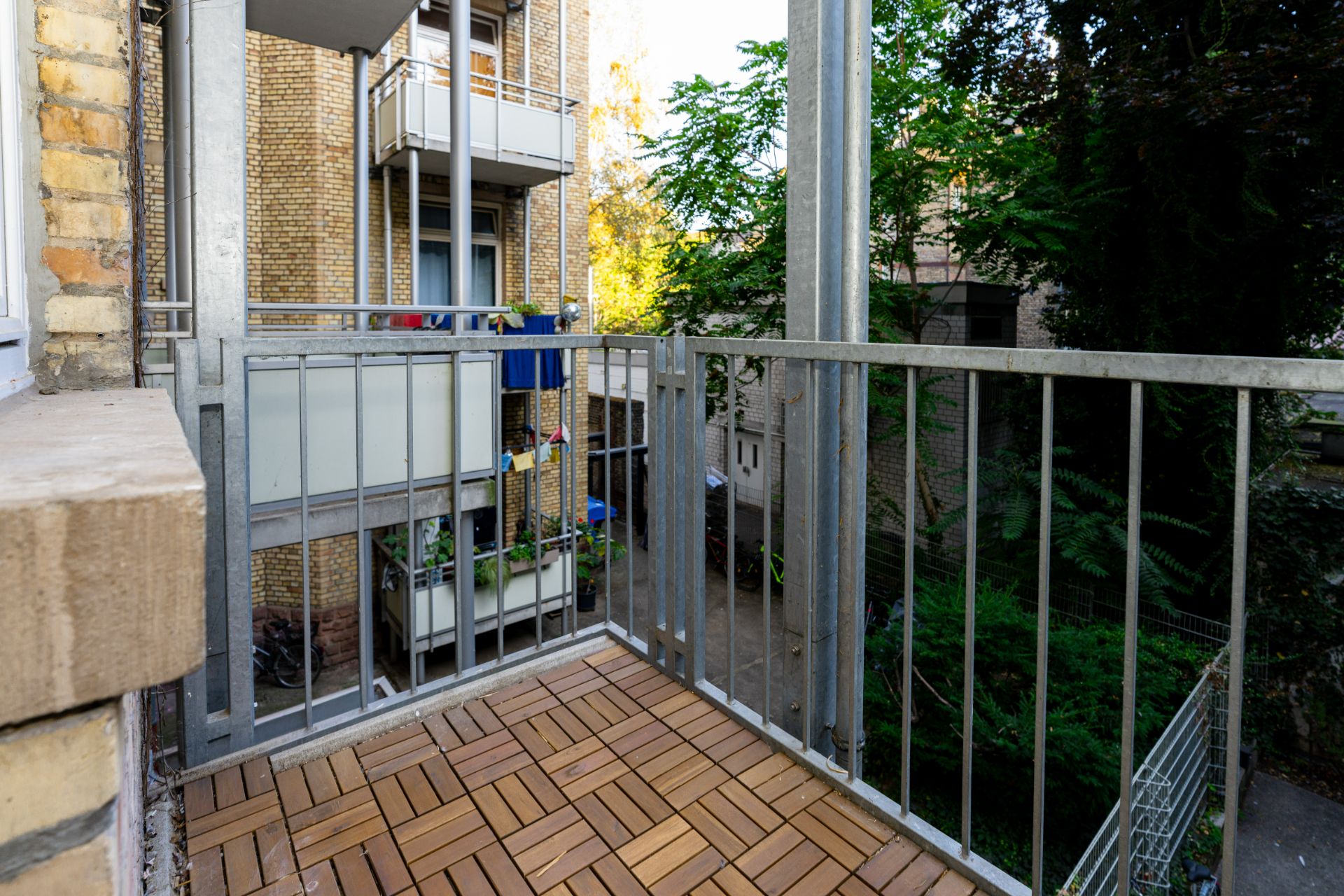 Kleiner Balkon mit Holzboden und Metallgeländer, Blick auf andere Balkone und grüne Bäume.