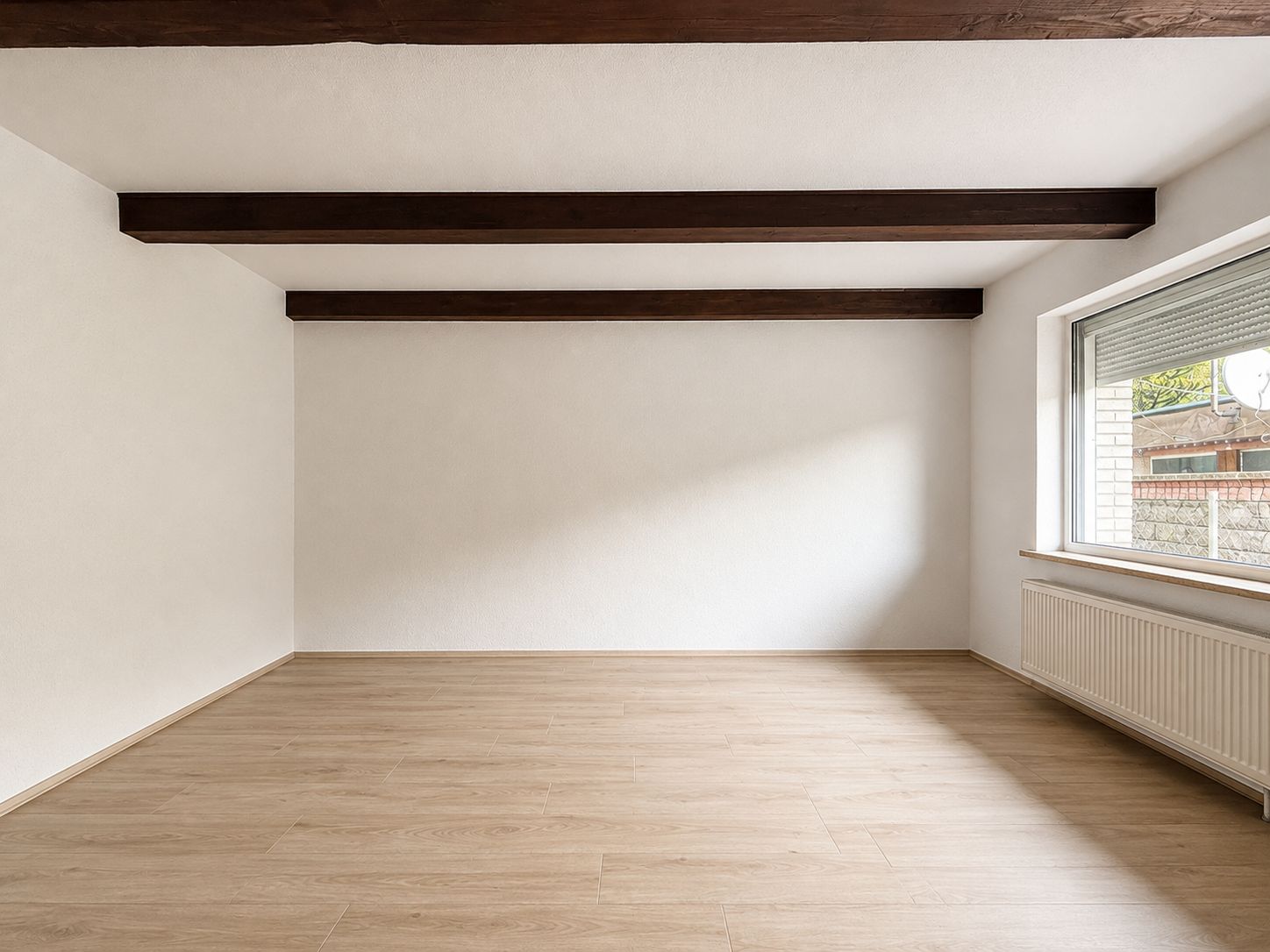 Leerer Raum mit hellem Holzboden, weißen Wänden, Holzbalken an der Decke und großem Fenster.