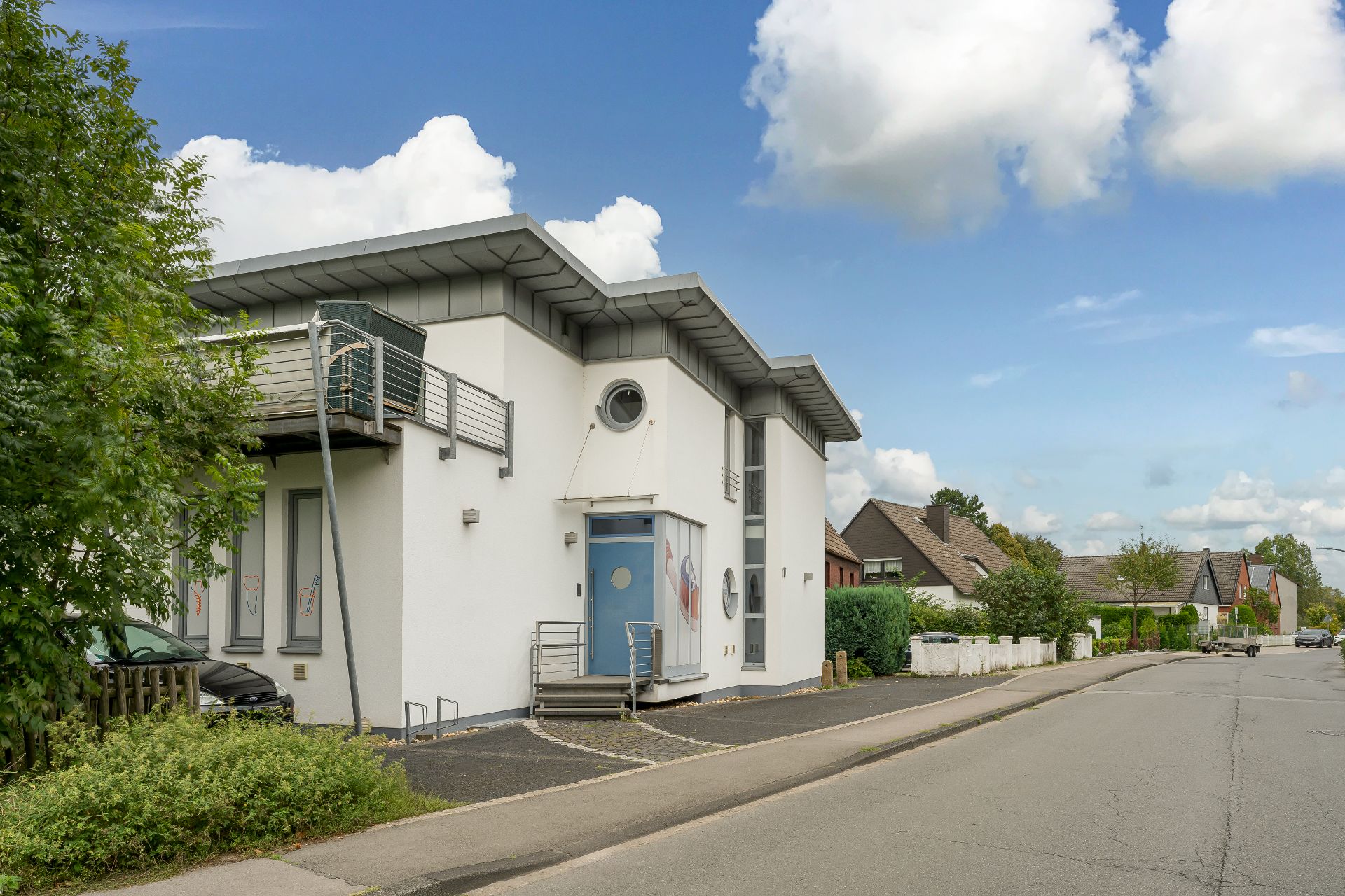Modernes zweistöckiges Wohnhaus mit großem Fenster und blauem Eingang an einer ruhigen Wohnstraße.